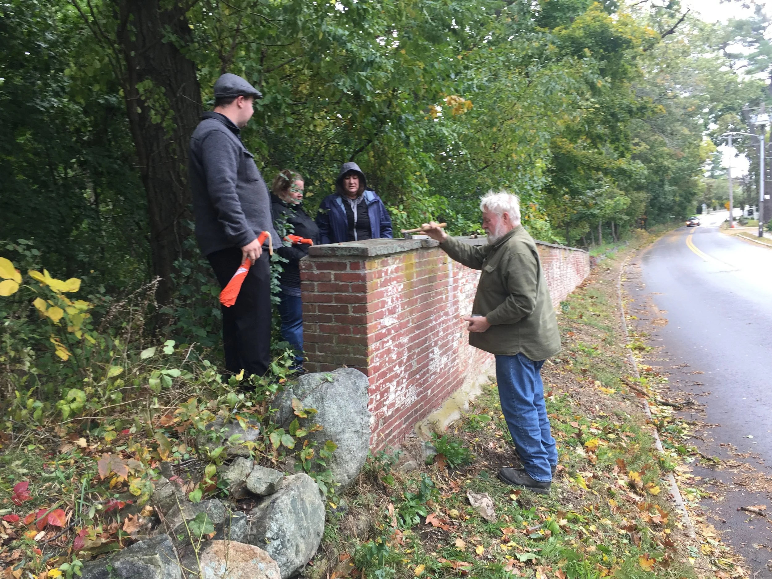 Group of people inspecting a brick wall near a wooded area and road.