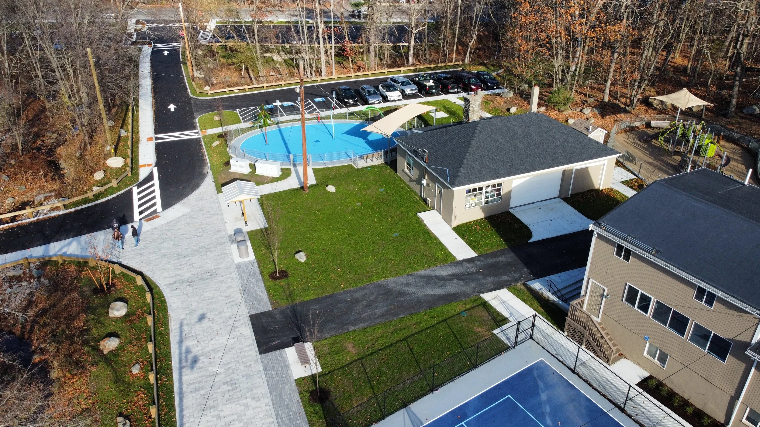 Aerial view of a recreational area with a pool, playground, tennis court, and parking lot, surrounded by trees.