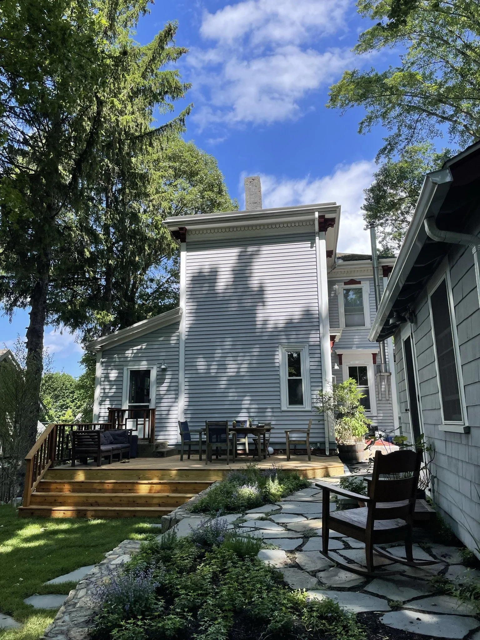 A light gray, two-story house with a wooden deck featuring outdoor furniture, surrounded by trees and greenery. There's a stone path leading to the deck and a wooden rocking chair in the foreground. The sky is blue with scattered clouds.