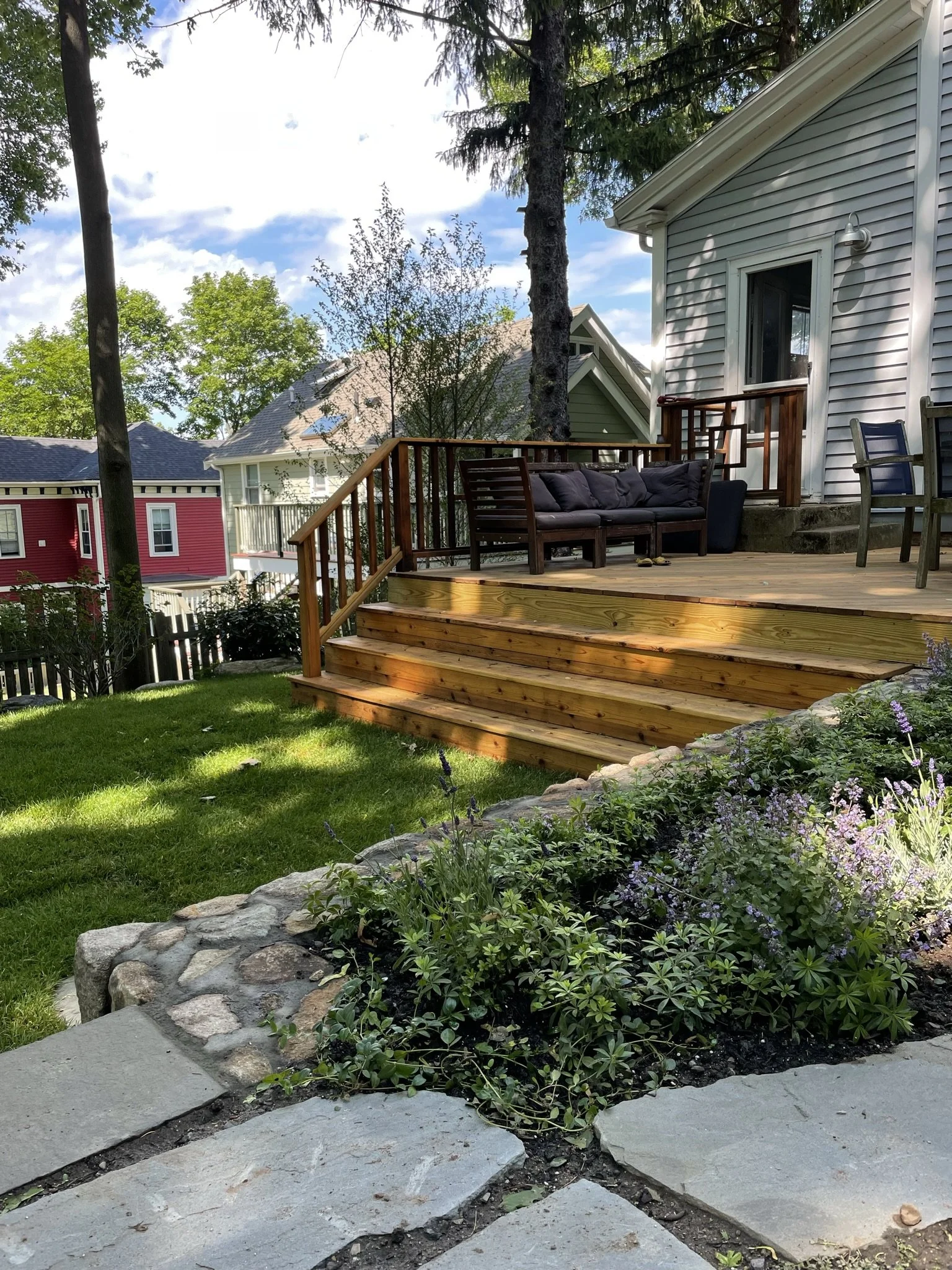 Backyard deck with wooden railing and outdoor furniture, surrounded by a lush garden and trees, with neighboring houses visible in the background.