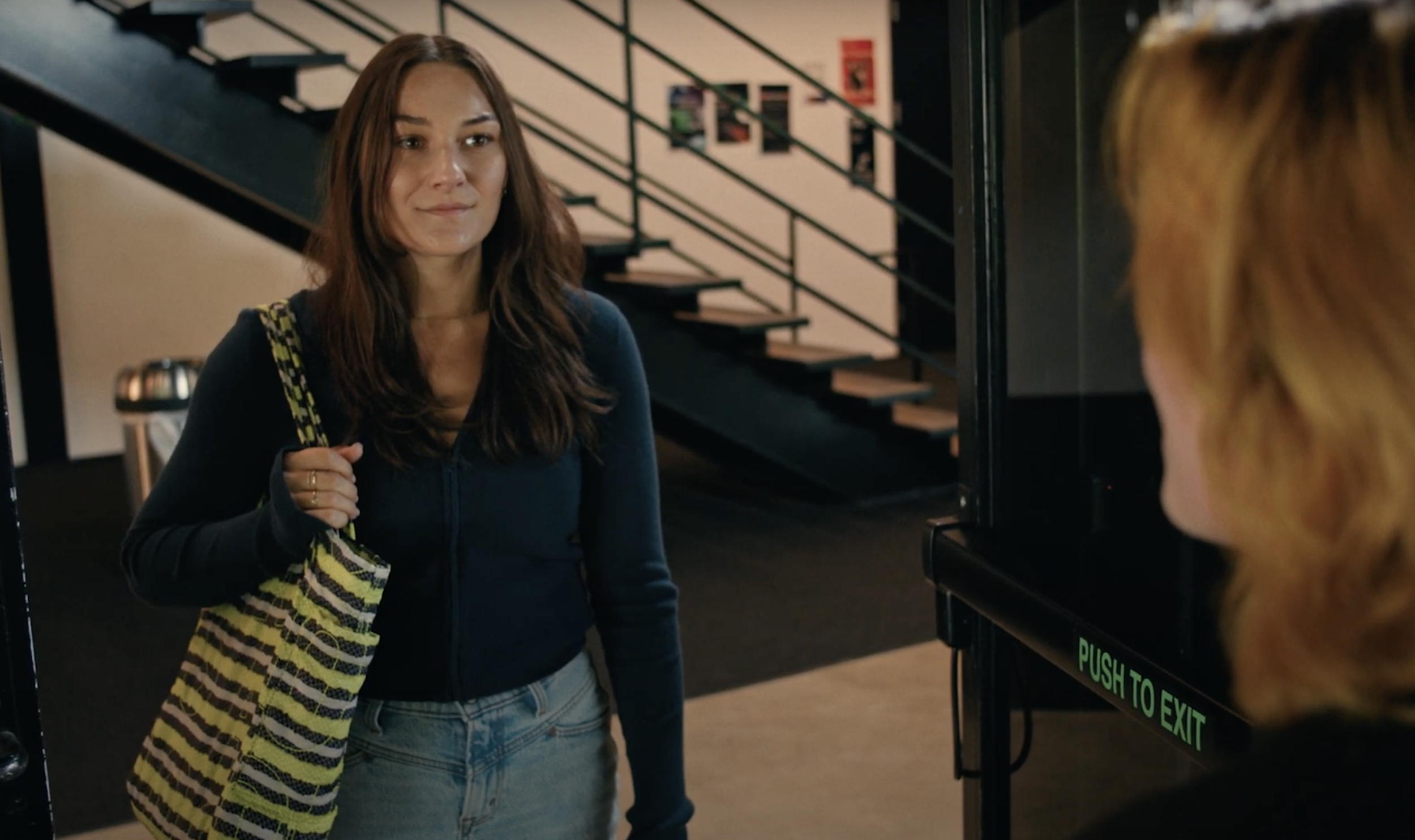 A still shot from a short film called "All Set” that Abby Marcus acting in. She sports a dark zip-up hoodie and a yellow and blue striped tote.