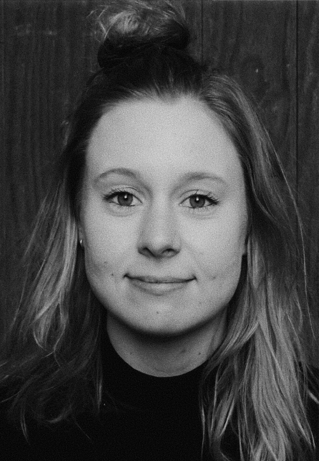 Black and white portrait of a woman with long wavy hair, wearing a dark top, and standing in front of a wooden background.