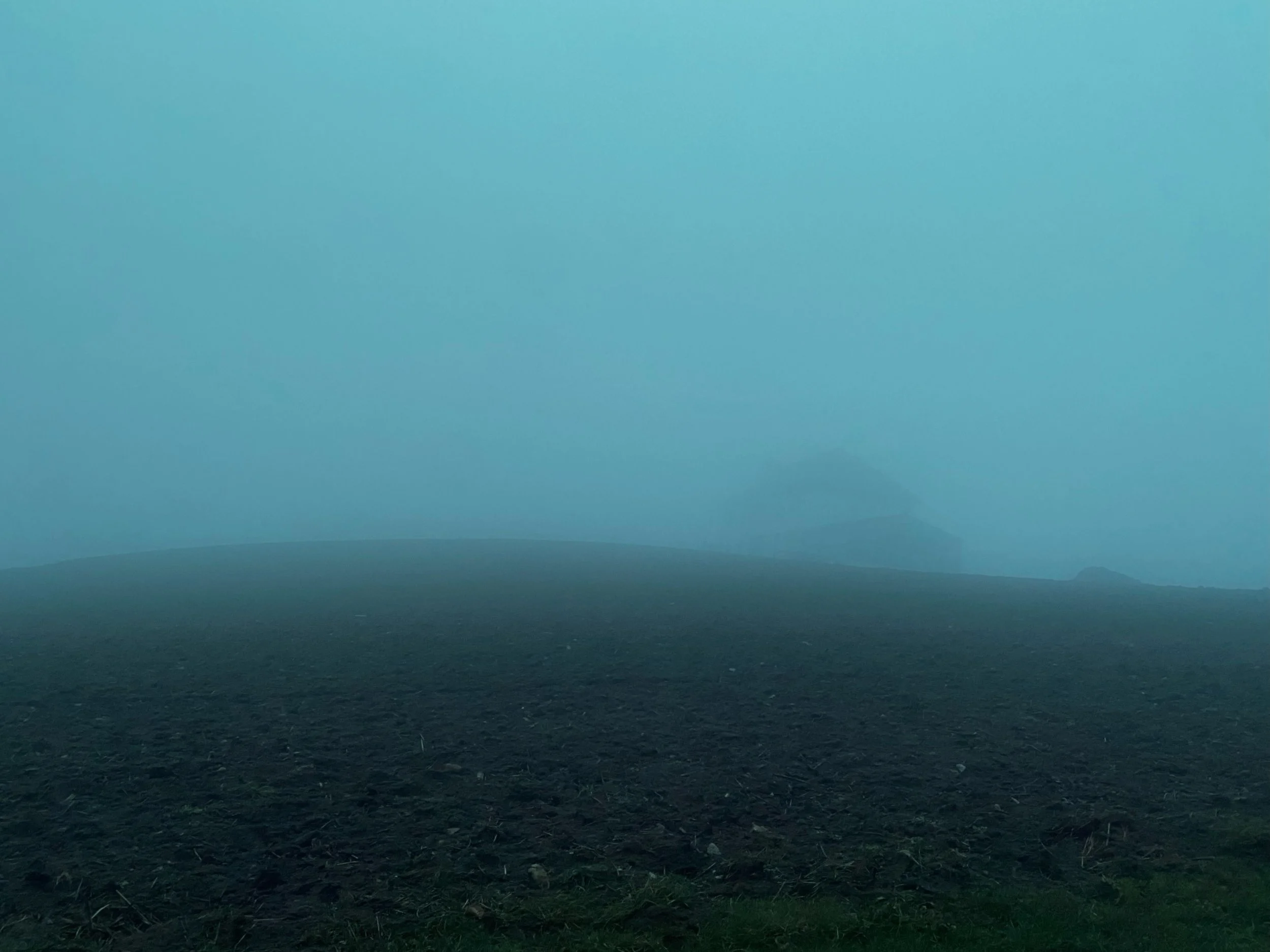 A foggy landscape with a dark, freshly plowed field in the foreground and misty hills in the background.