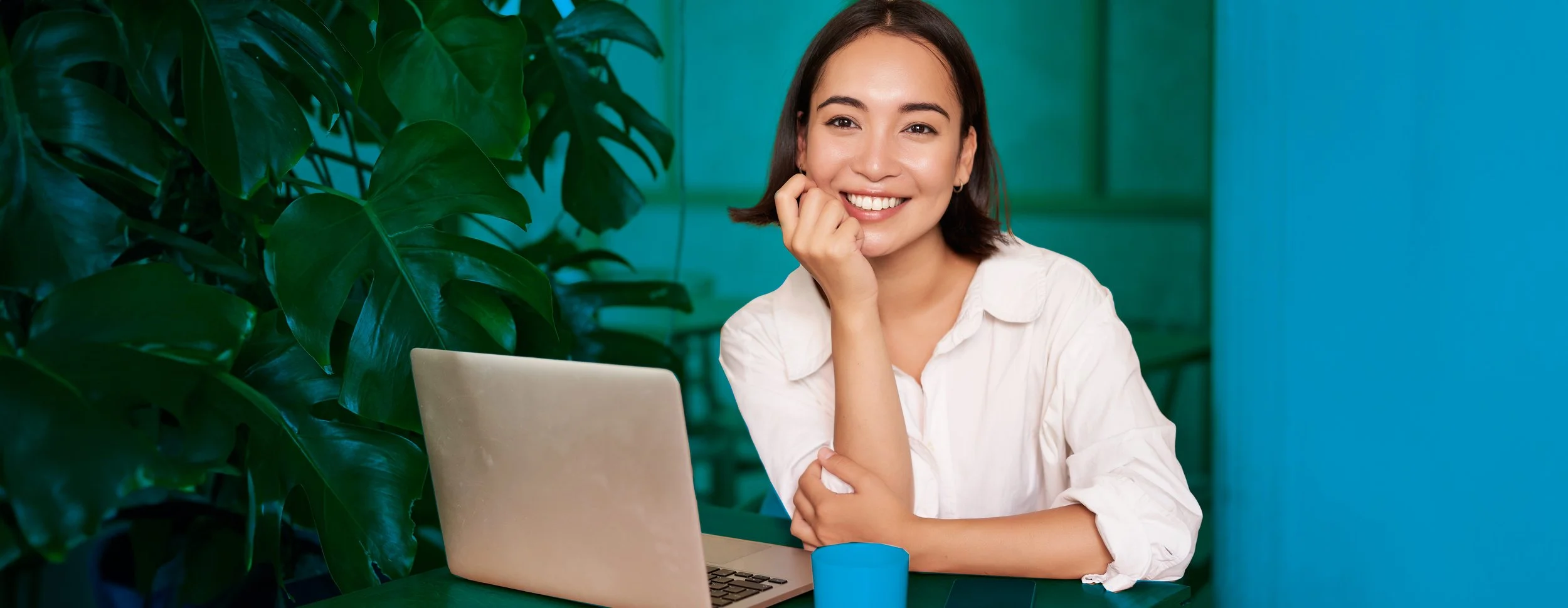 Joven mujer sonriendo, sentada en una mesa con una computadora portátil y una taza azul, rodeada de plantas verdes.