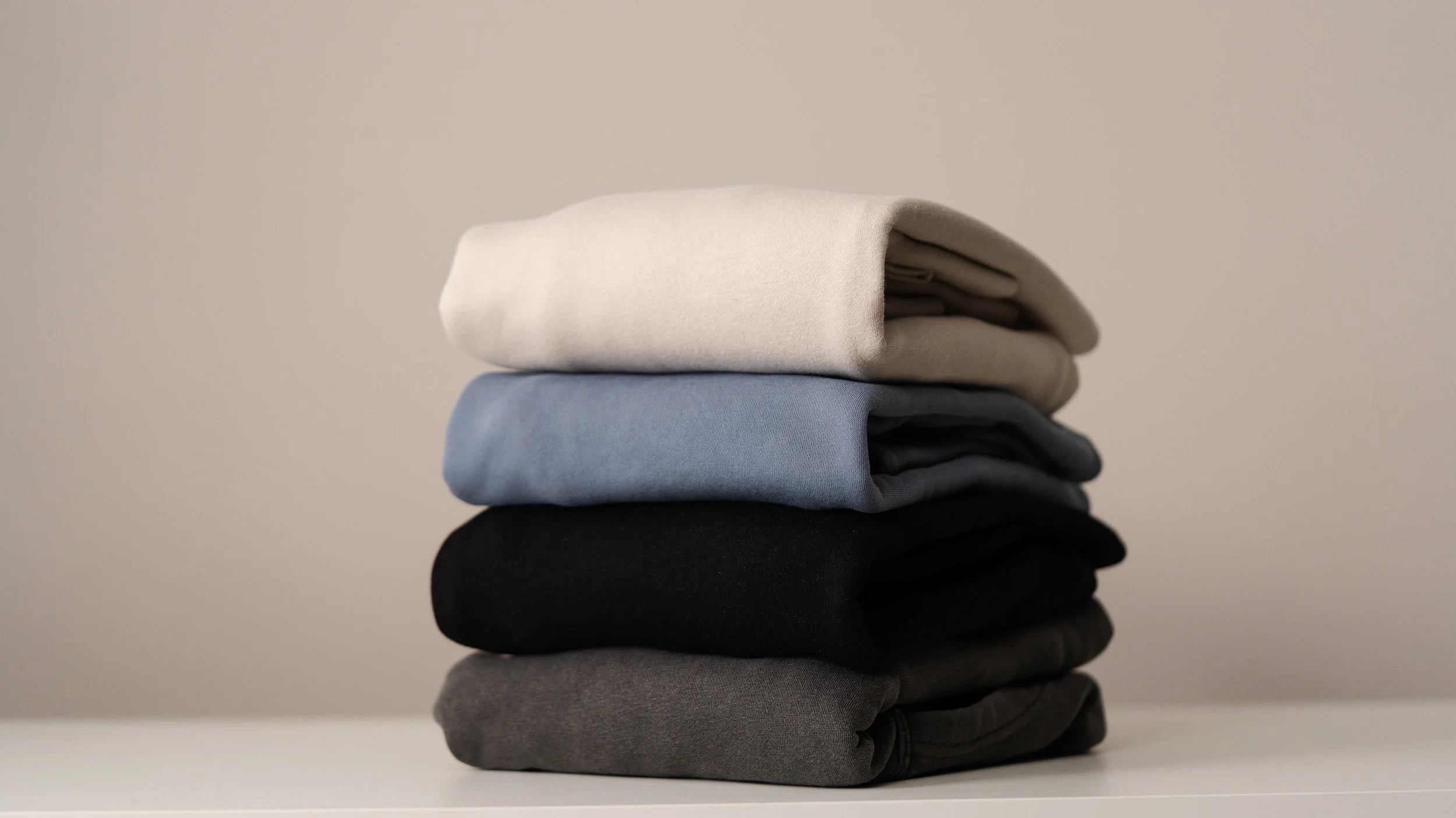 A neatly folded stack of five pairs of pants or trousers in different colors including beige, light blue, black, and gray, on a white flat surface against a plain light background.