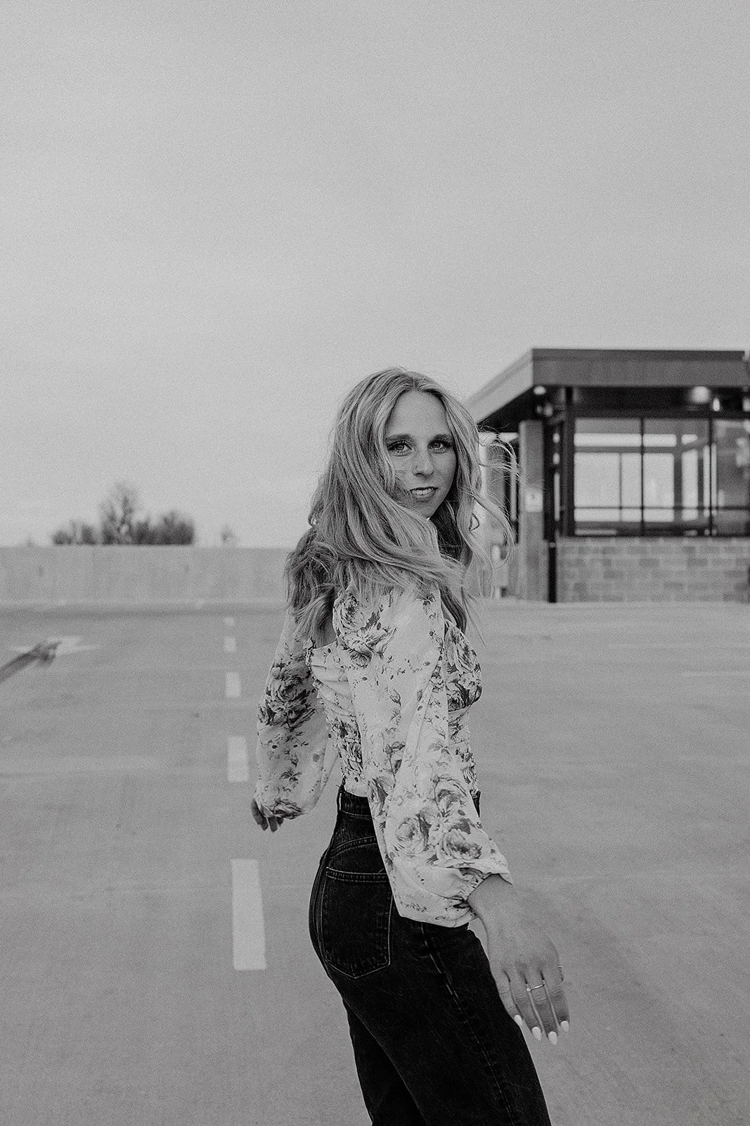 A woman with long, curly hair stands on an empty rooftop parking lot with a building in the background. She is wearing a floral long-sleeve top and dark jeans, looking over her shoulder at the camera.