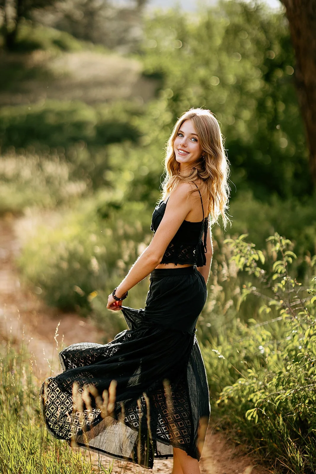 A young woman in a black lace dress standing outdoors in a green, wooded area, smiling at the camera with sunlight filtering through the trees.