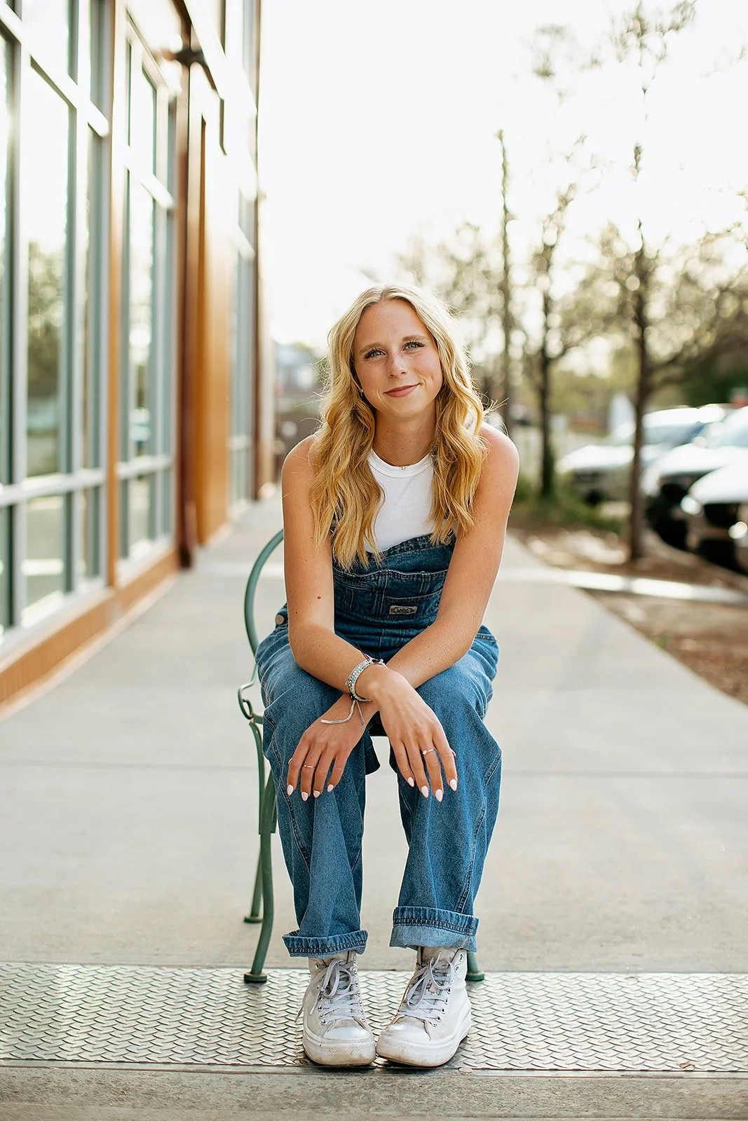 A young woman with blonde wavy hair smiling while sitting on a metal bench outside a modern building with large glass windows; she is wearing a white sleeveless top, blue denim overalls, and white sneakers, with jewelry including rings, bracelets, an