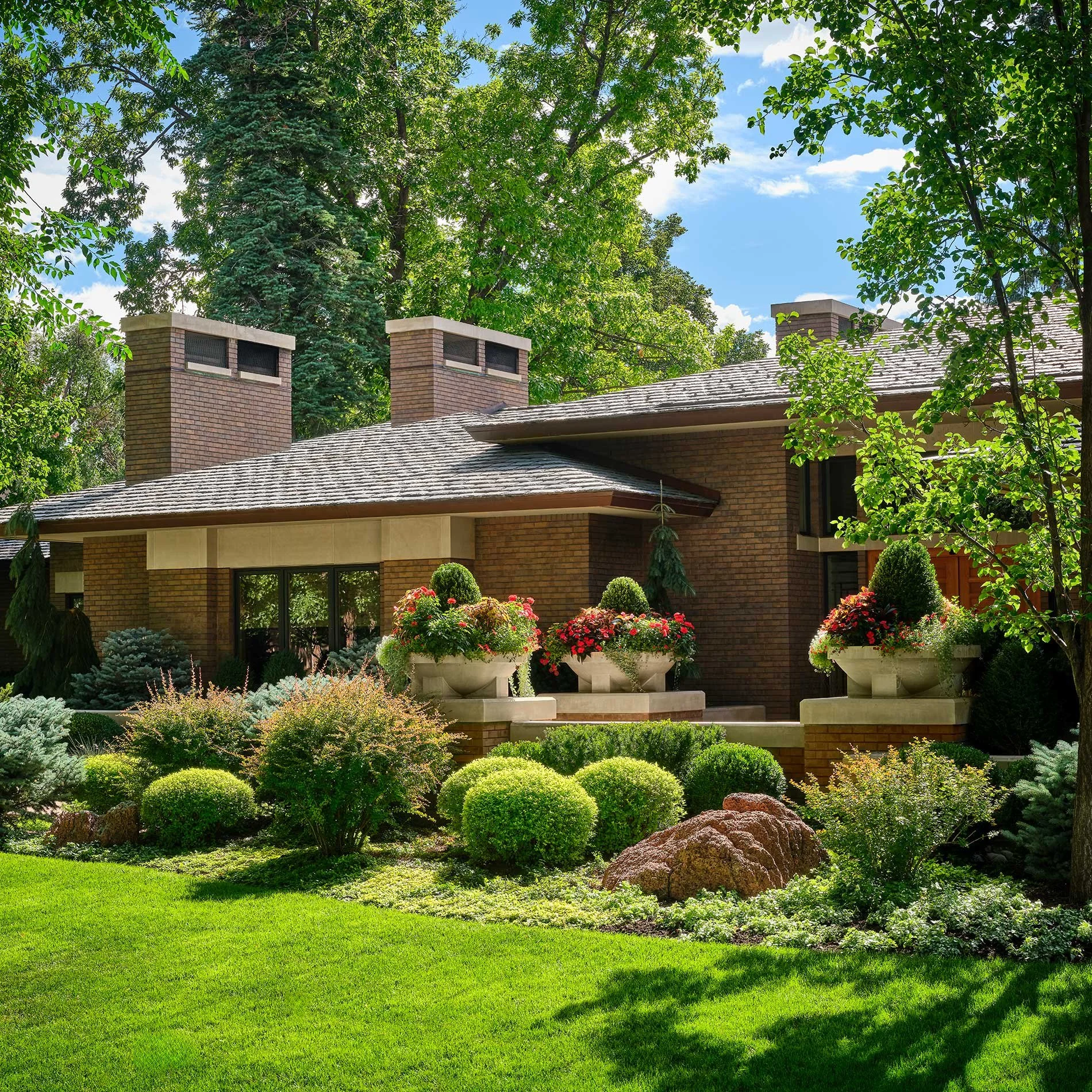 A brick house with a sloped roof surrounded by lush green trees and well-maintained garden with various shrubs, flowers, and a large rock.