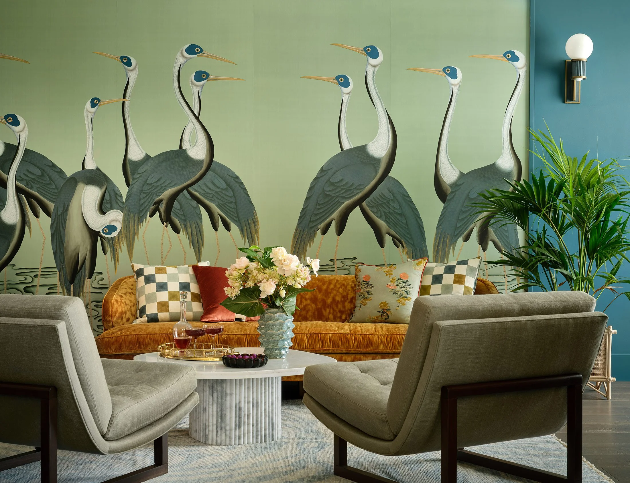 Living room with a mural of large usian cranes on the wall, a mustard-colored velvet sofa with decorative pillows, a round marble coffee table with a flower vase and drinks, and two gray armchairs. There is a green plant and a wall-mounted light.
