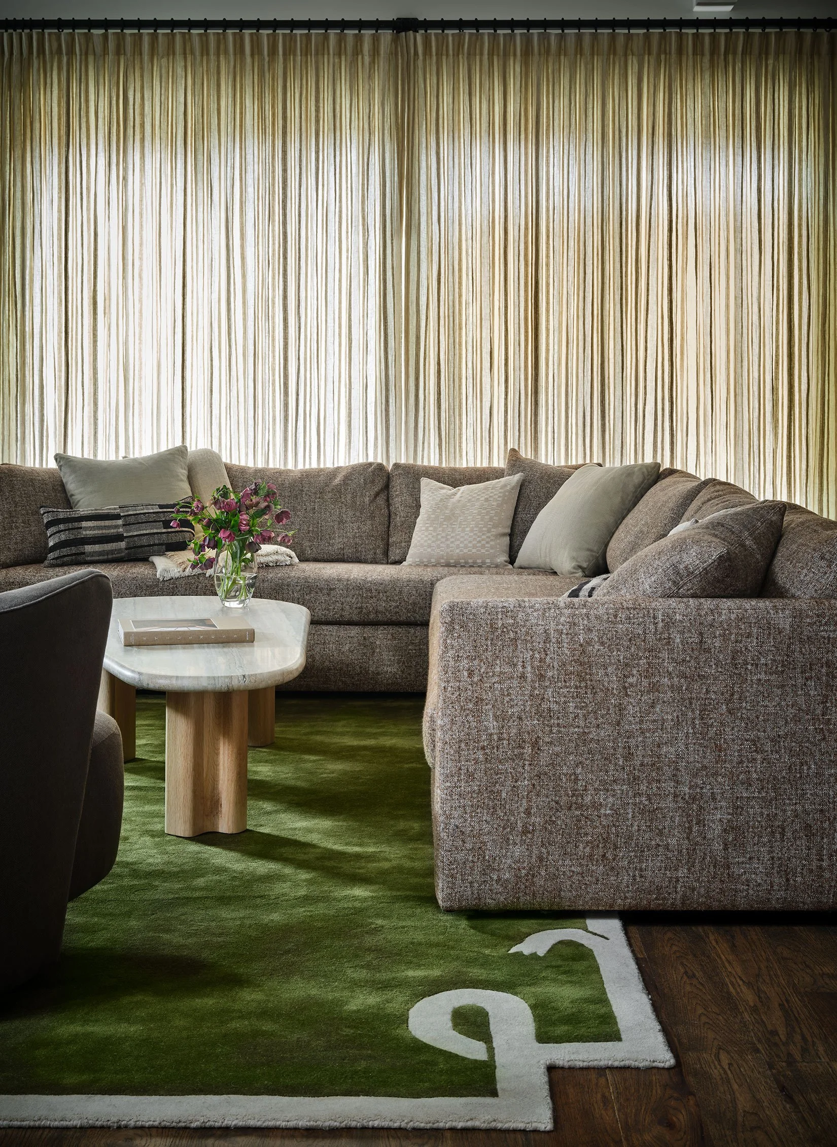 Living room with a large beige sectional sofa, decorative pillows, a marble coffee table with a vase of flowers, and beige curtains covering a window, with a green area rug on wooden floor.