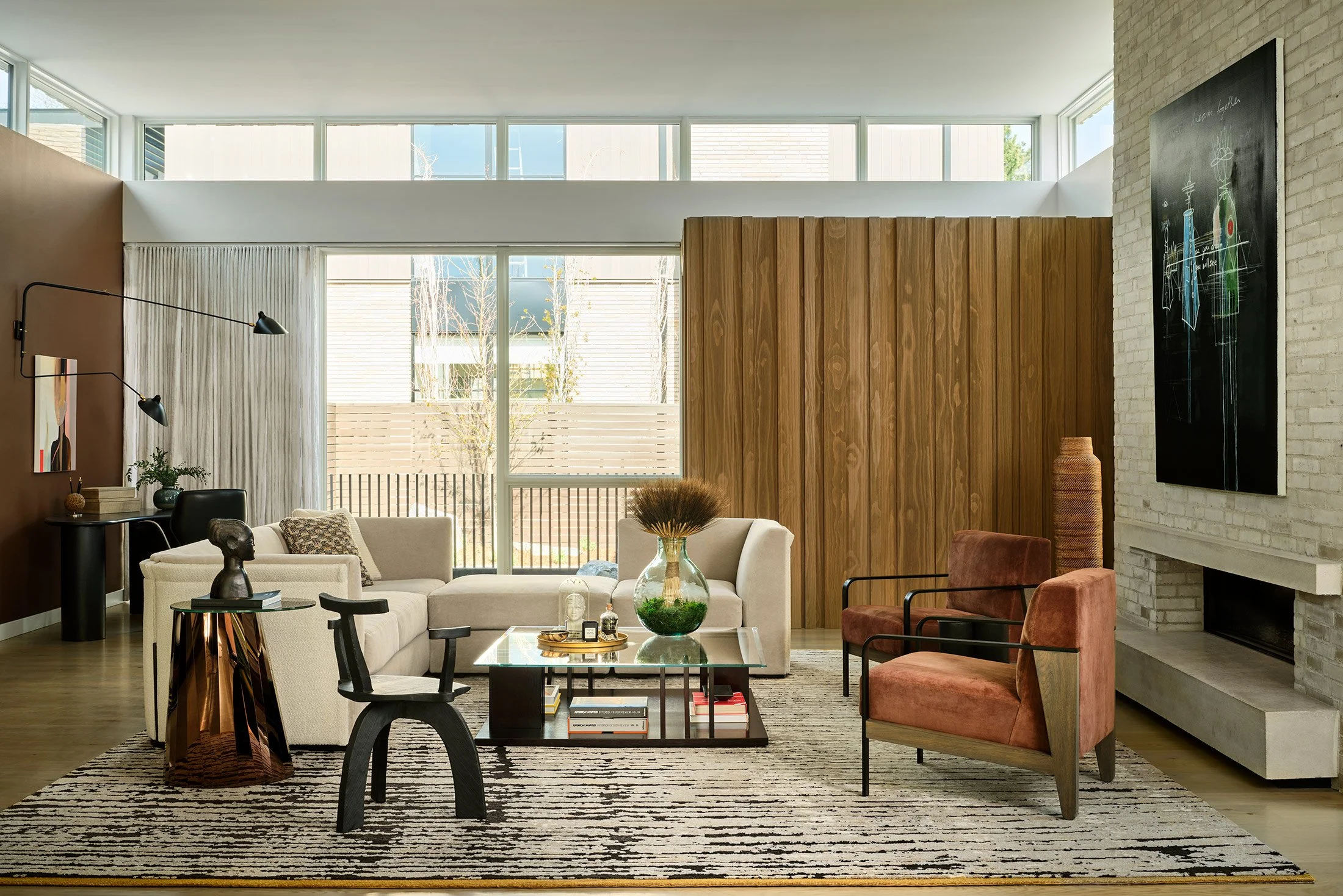 Modern living room with beige sectional sofa, two rusty orange armchairs, a black side table, and a glass coffee table with books and decorative items. Large windows, wooden panel wall, white brick fireplace, and artwork on the walls.