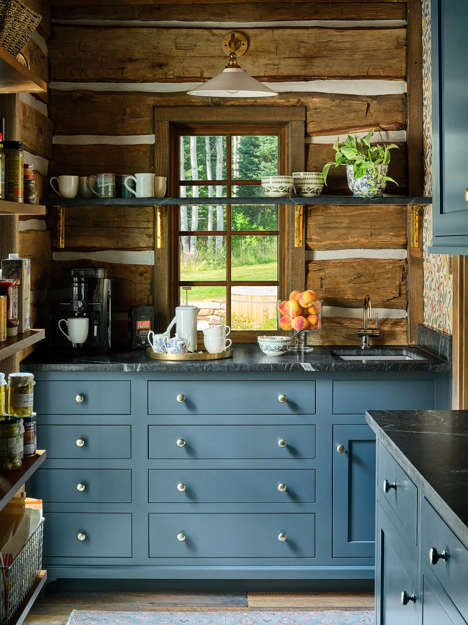 A rustic kitchen with blue cabinets, black countertops, wooden walls, and a window showing an outdoor green landscape. The countertop has a coffee maker, teapots, cups, a bowl of peaches, and a potted plant. Shelves hold bowls and mugs.