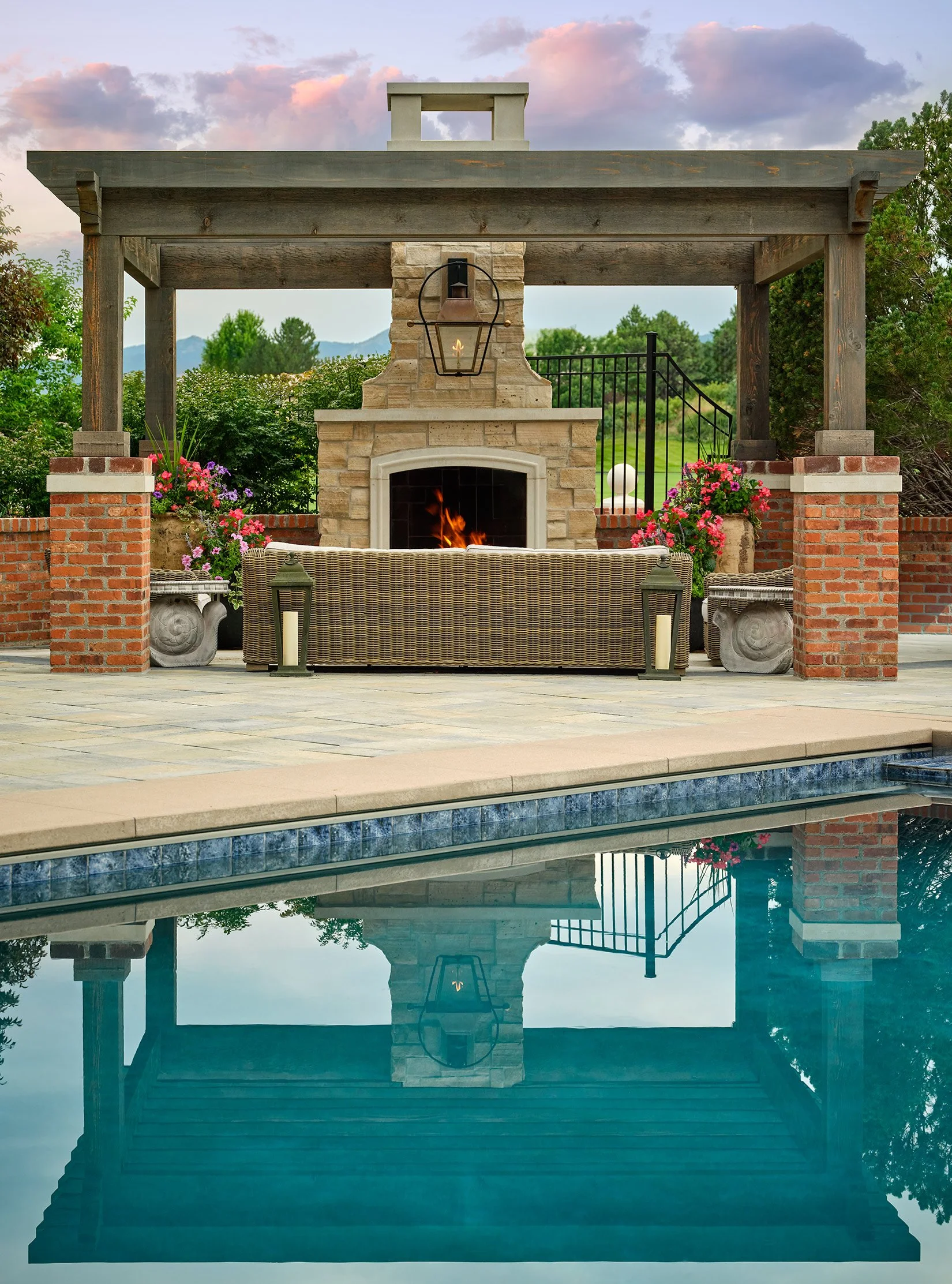 Outdoor patio area with a swimming pool in the foreground, featuring a stone fireplace with a chimney, a lantern hanging above it, a wicker couch, and flower pots with pink and purple flowers on brick pillars. The scene is set during sunset with a co