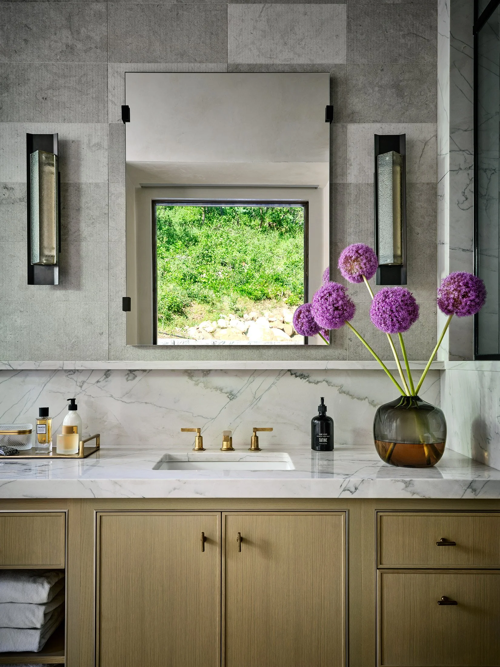 Modern bathroom with beige and grey cabinetry, marble countertop, gold fixtures, a large mirror, and a vases with purple flowers. Outdoors green landscape visible through window.