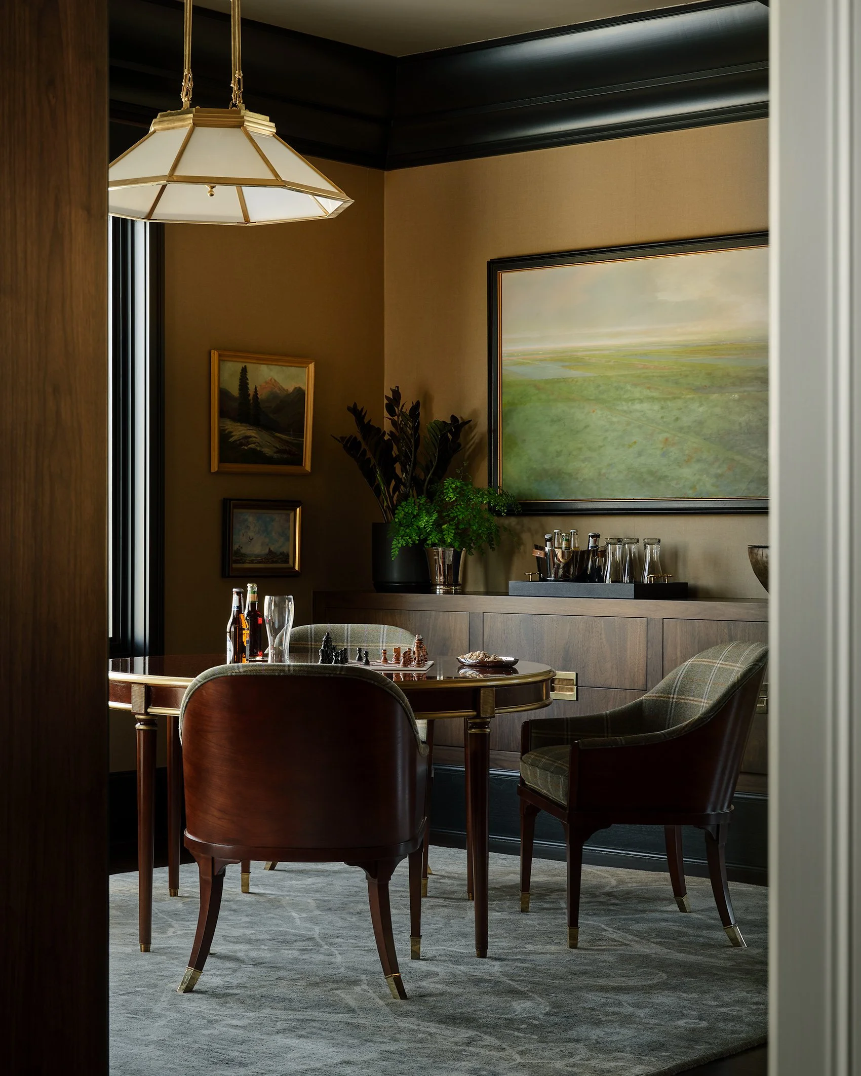 An elegant meeting room with a round wooden table, four chairs, a large landscape painting, smaller framed artwork, a industrial style pendant light, a side cabinet with glasses and bottles, and a plant, with dark paneled walls and window with black 