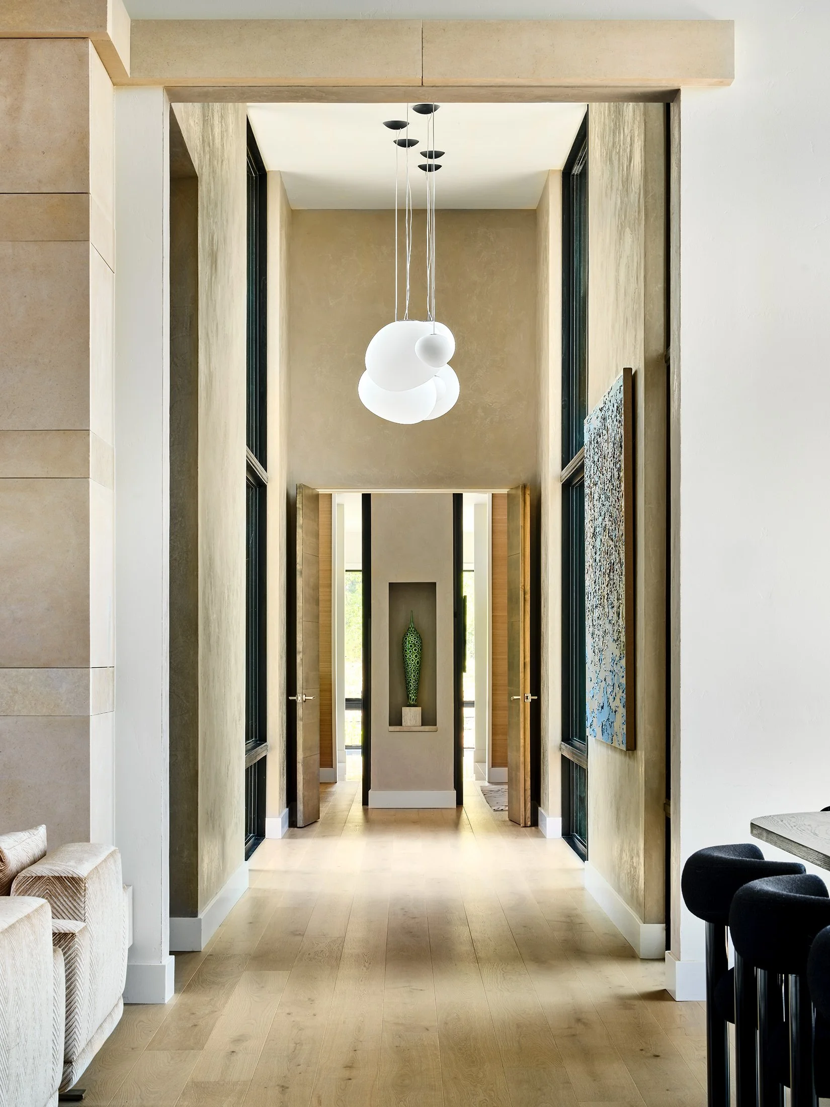 Modern interior hallway with high ceilings, hanging white globe pendant lights, beige walls, natural wood flooring, a decorative vase in a niche, and artwork on the wall.