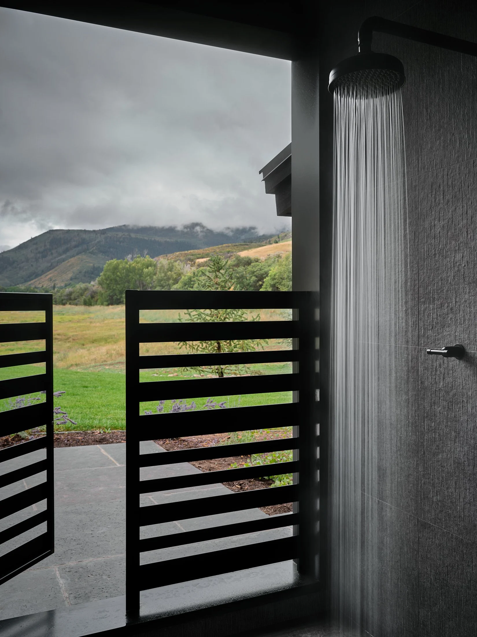 Outdoor shower with rainfall head on dark textured wall, overlooking mountains, cloudy sky, and lush greenery.