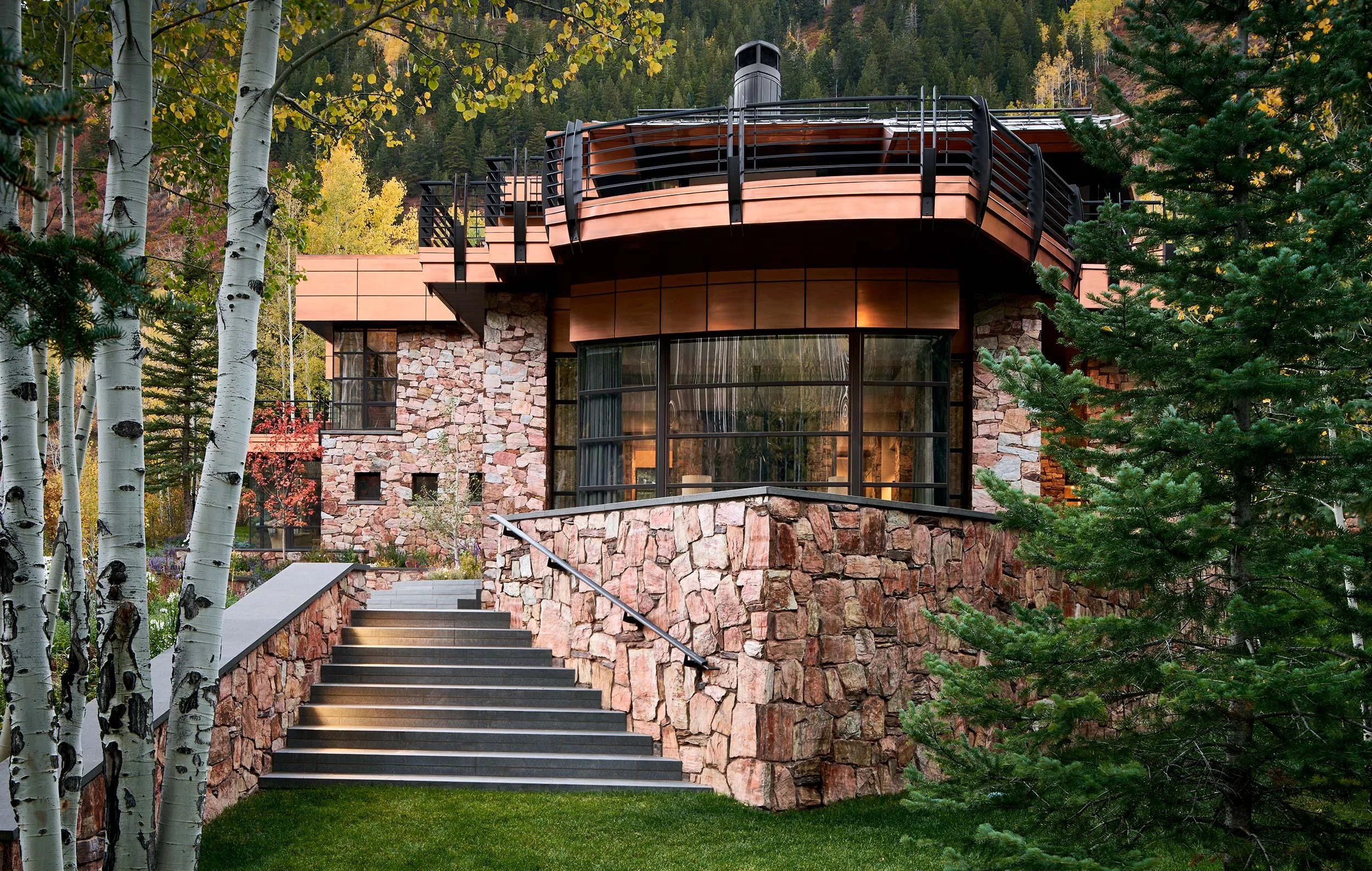 A modern house built with stone, glass, and wood, situated in a forested area with trees and colorful autumn leaves in the background.