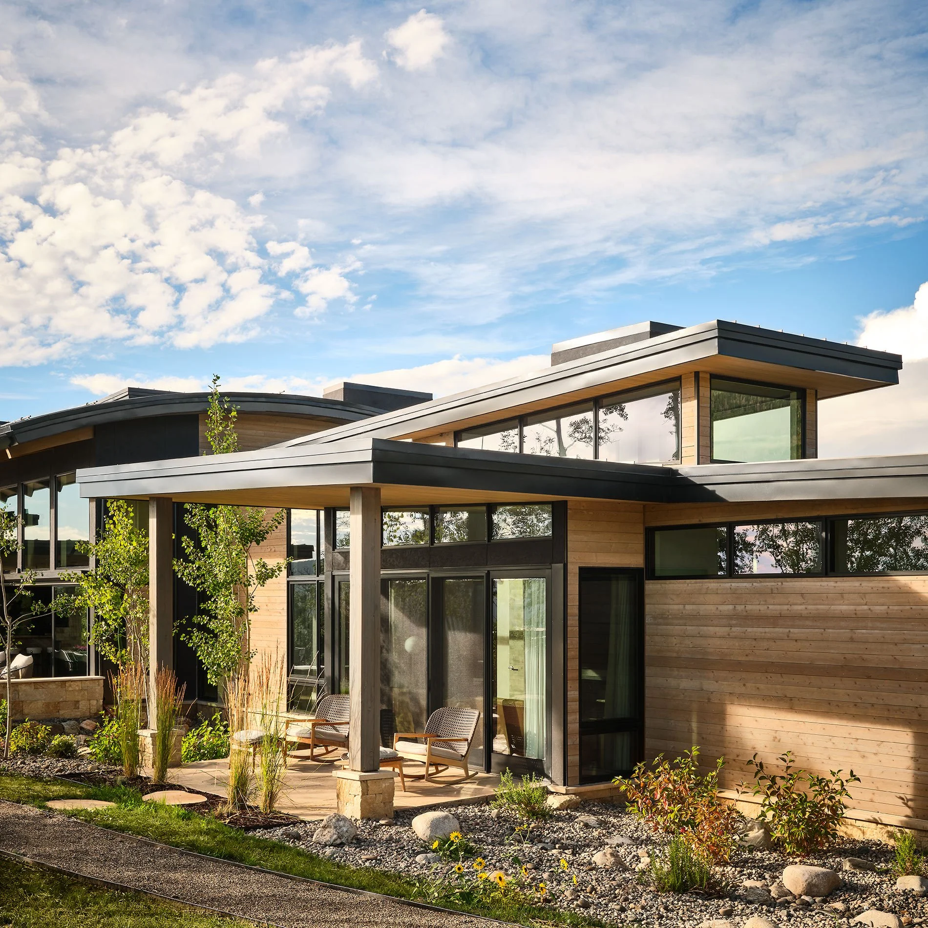 Modern house with large glass windows and a small outdoor sitting area with chairs, surrounded by plants and rocks under a partly cloudy sky.