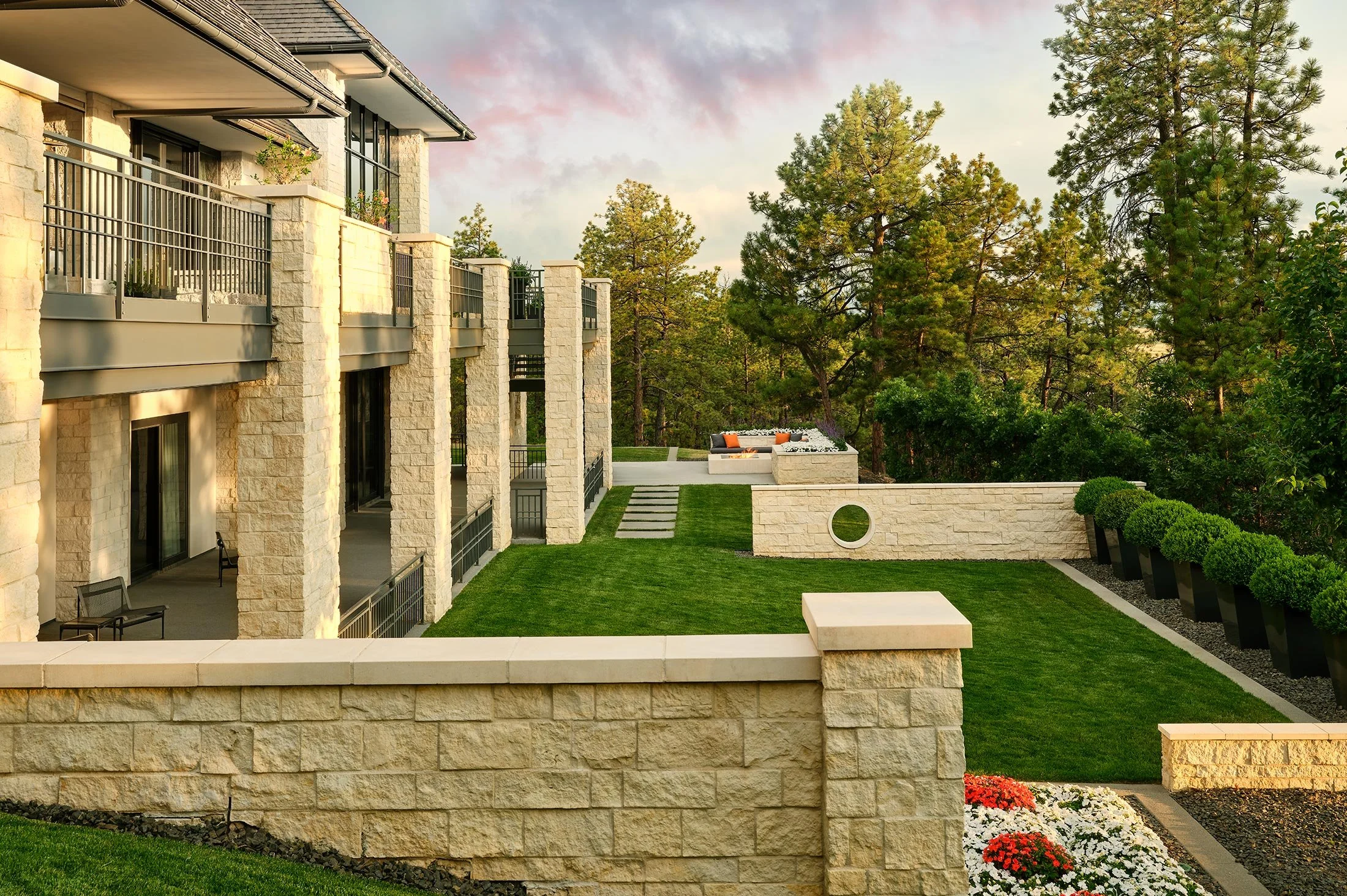 Modern residential building with beige stone walls, black metal railings, and a manicured lawn. There is an outdoor seating area with white cushions and orange pillows, surrounded by trees and shrubs. Flower beds with white and red flowers are in the