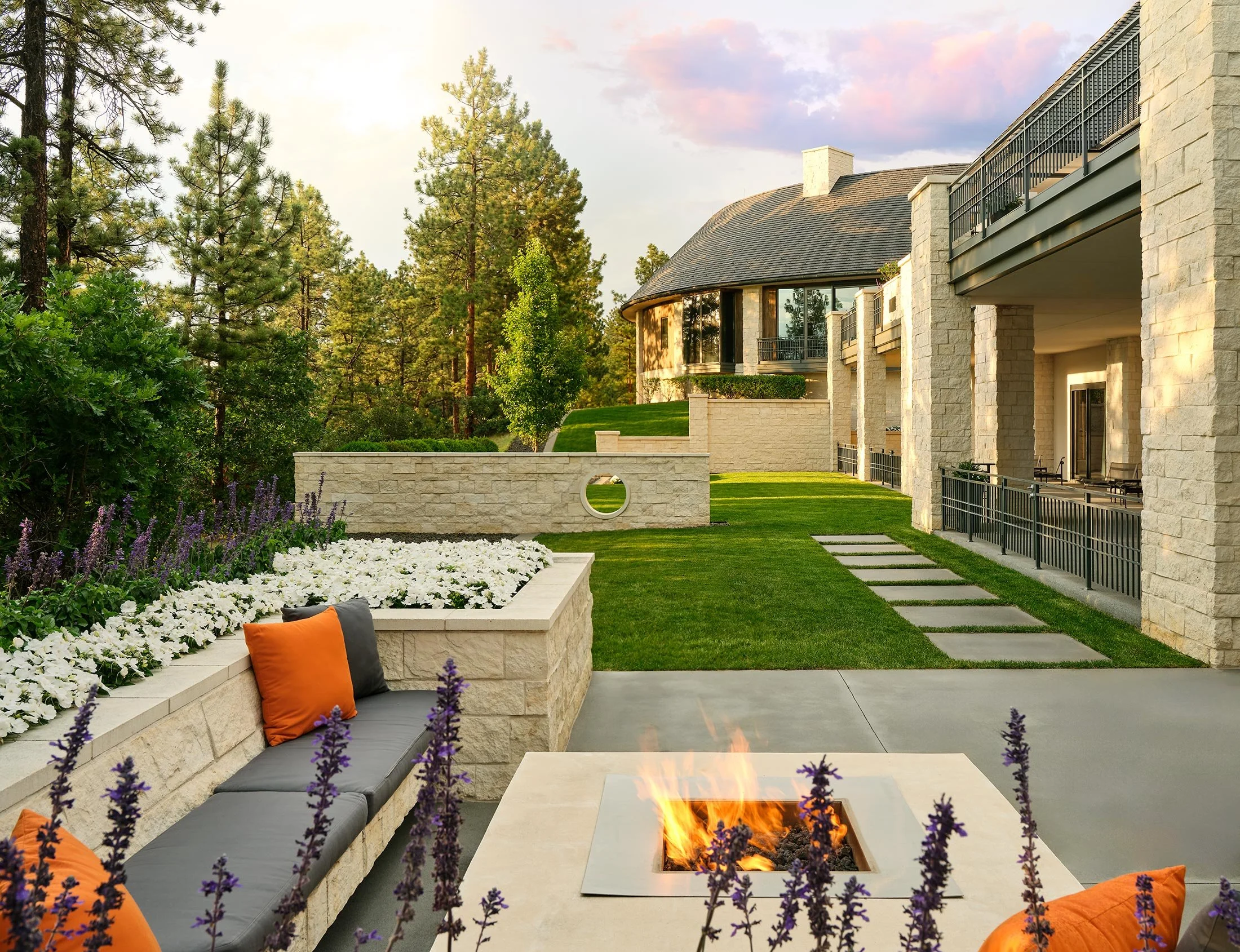 Spacious backyard patio with seating area, modern fire pit, colorful cushions, lush green lawn, garden beds with flowers, and a large house with stone walls and balconies, surrounded by tall trees and a sunset sky.