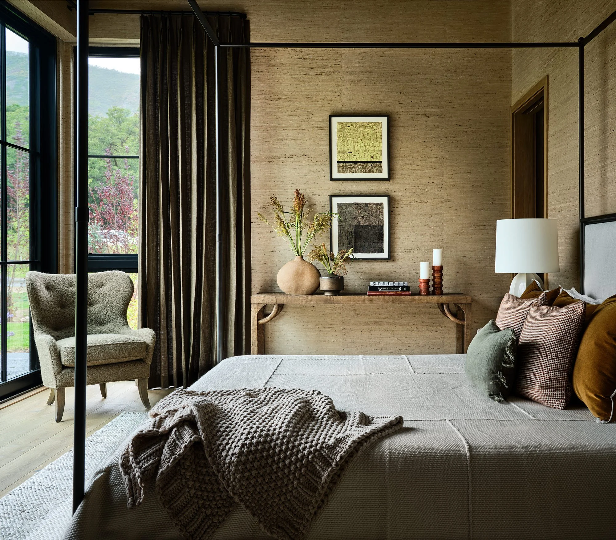 A cozy bedroom with a bed, decorative pillows, a lamp, and a wooden side table with vases, candles, and books. Large windows with dark curtains overlook a garden with trees.