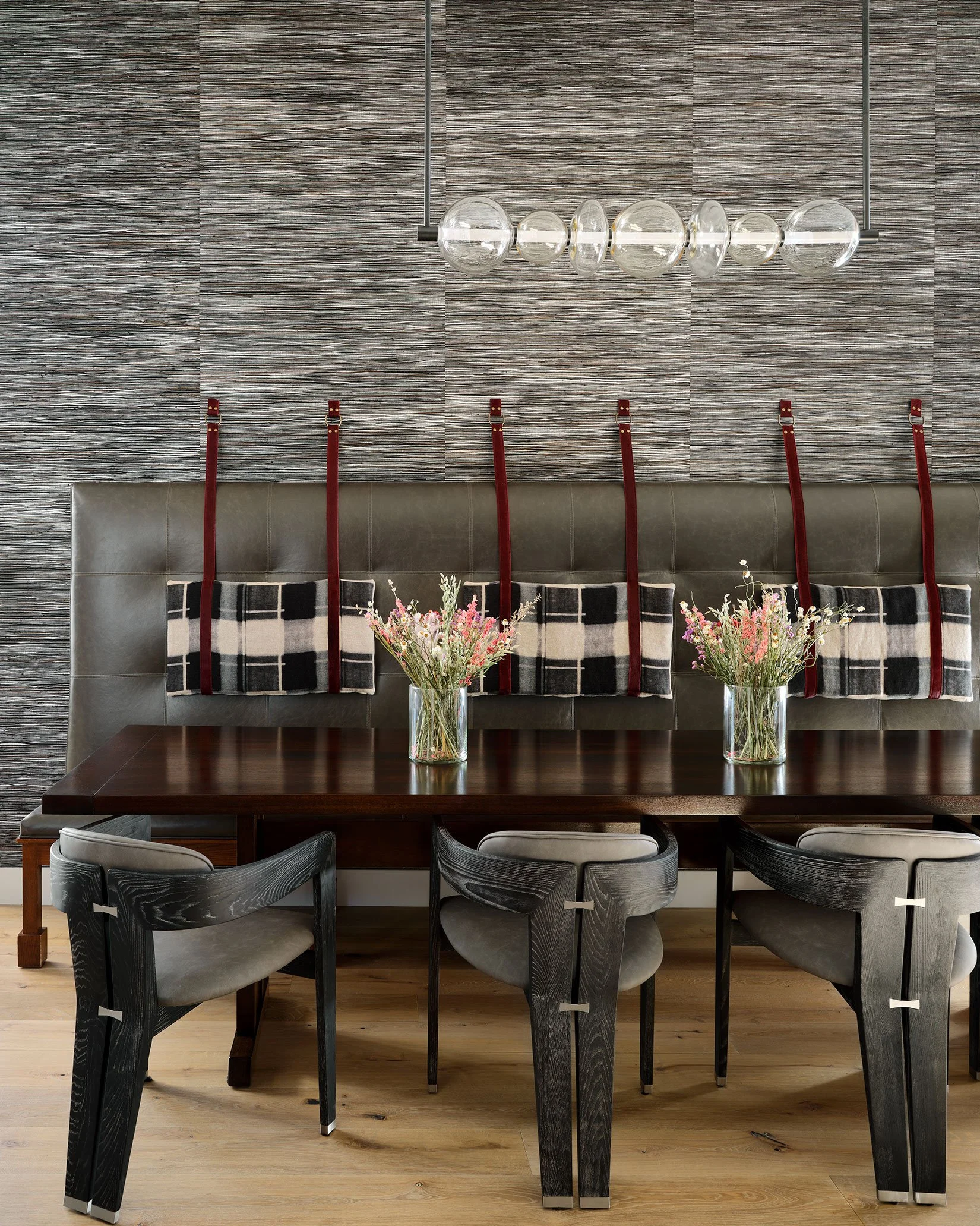 A modern dining area with a dark wooden table, black and white upholstered chairs, a gray cushioned bench with red straps attached to plaid pillows, glass vases with pink and white flowers, a textured wall, and a contemporary light fixture with glass