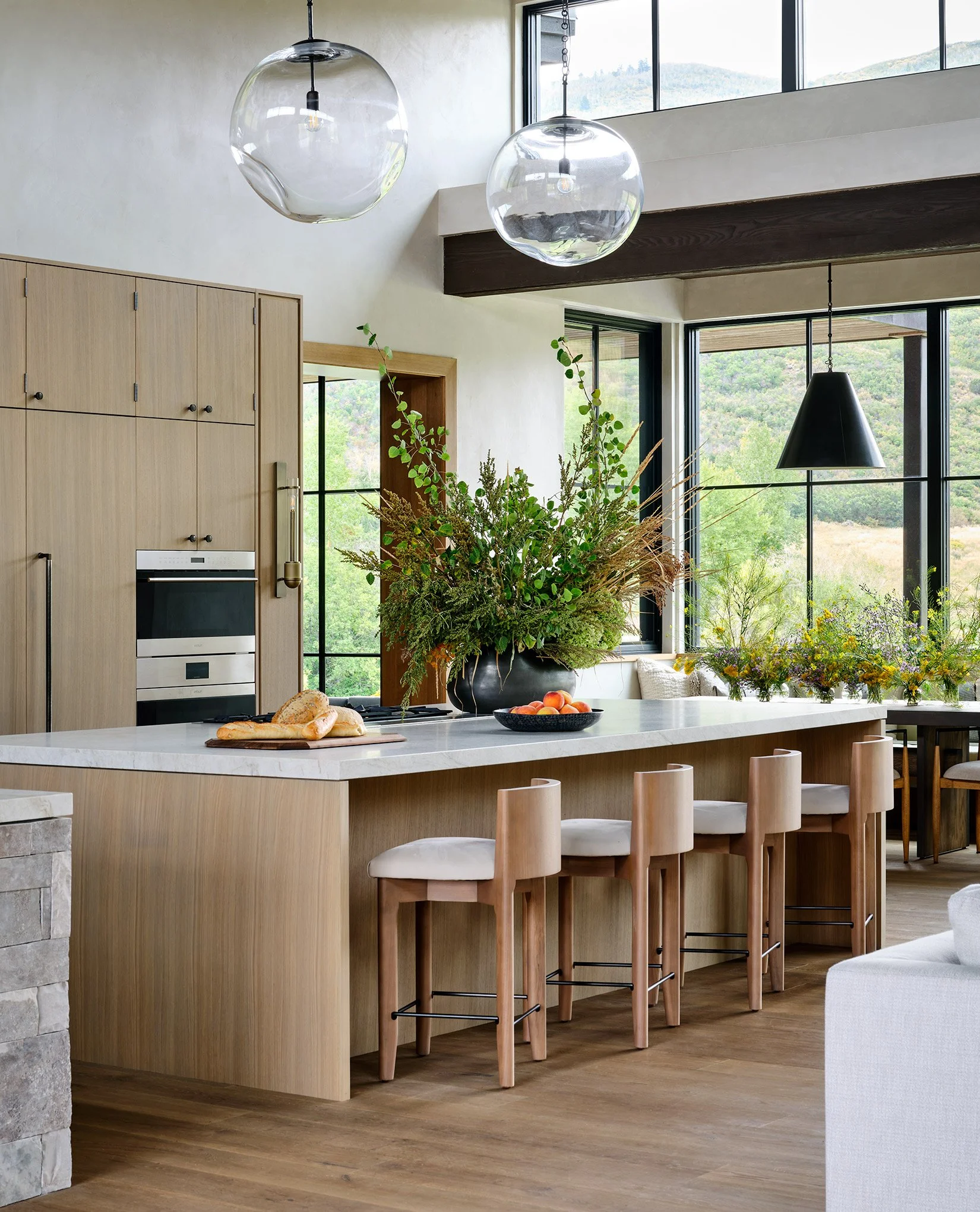 Modern kitchen with a large island, six wooden barstools, and a vase with greenery, located near large windows showing a scenic outdoor view.