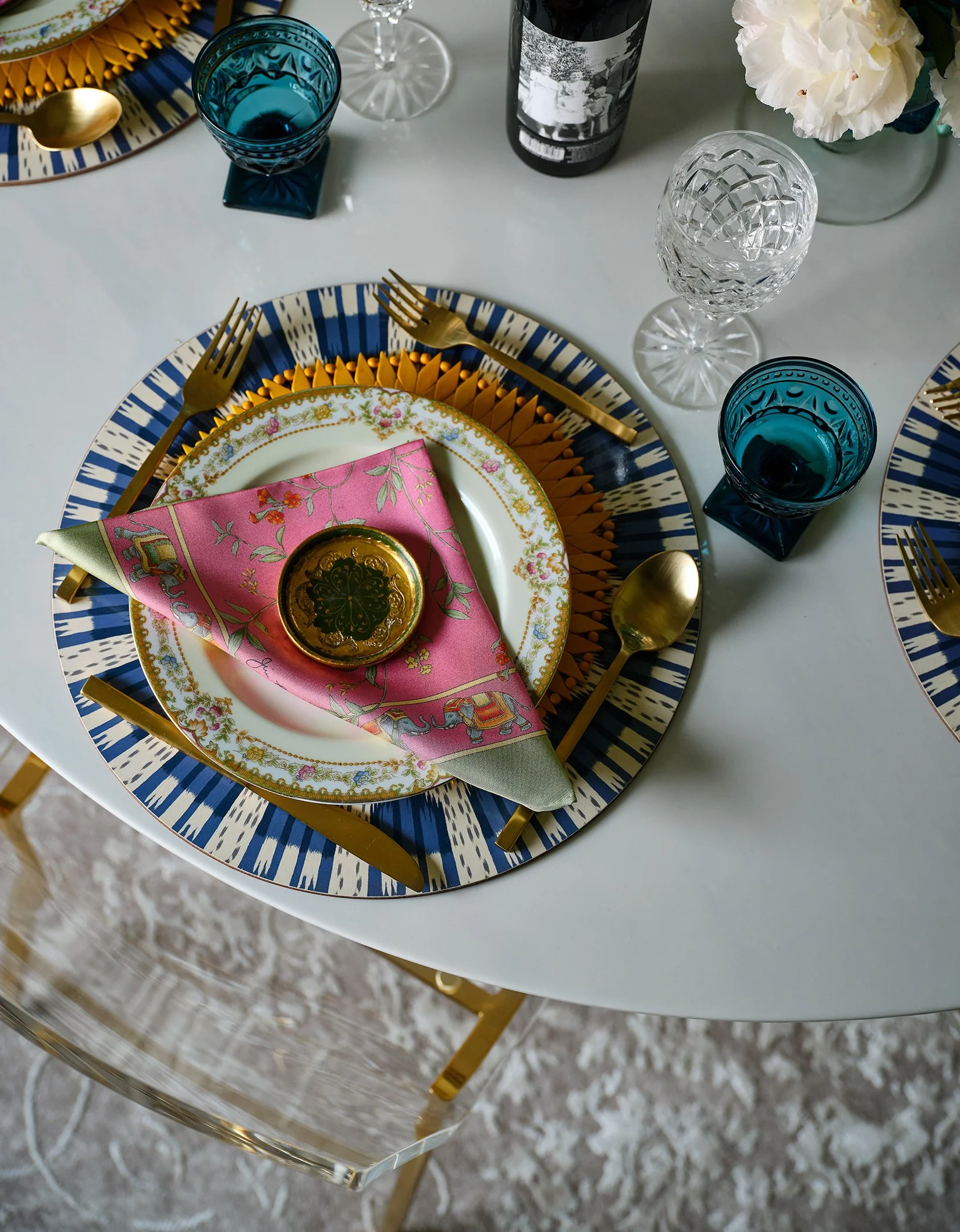 A table setting with blue and white patterned chargers, gold flatware, decorative plates, a pink floral napkin with a small gold dish, two blue glasses, a clear textured glass, a bottle of wine, and a flower arrangement.