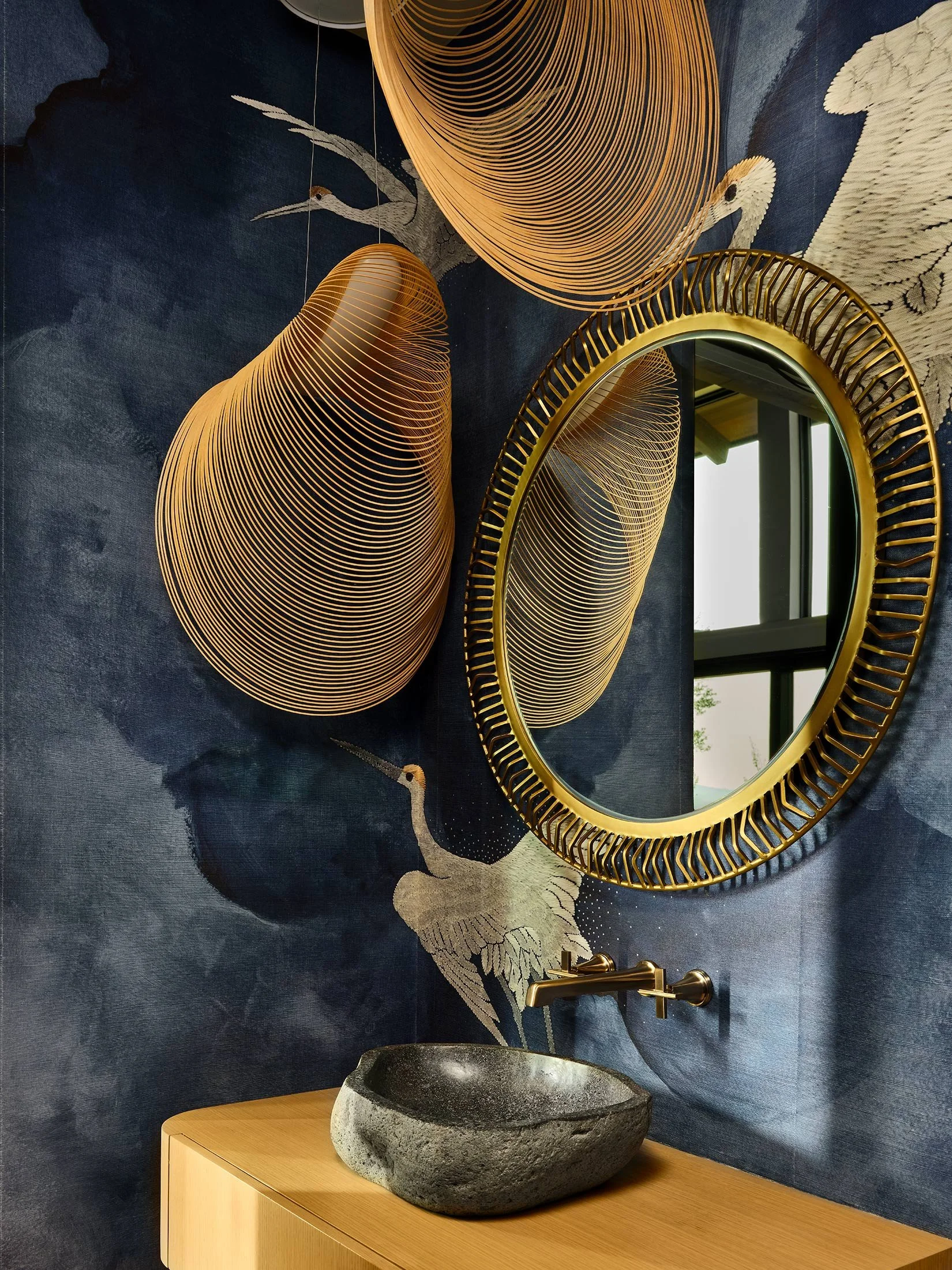 A bathroom with a round mirror, three hanging wooden pendant lights, a stone vessel sink, and a painted wall featuring cranes and abstract clouds.