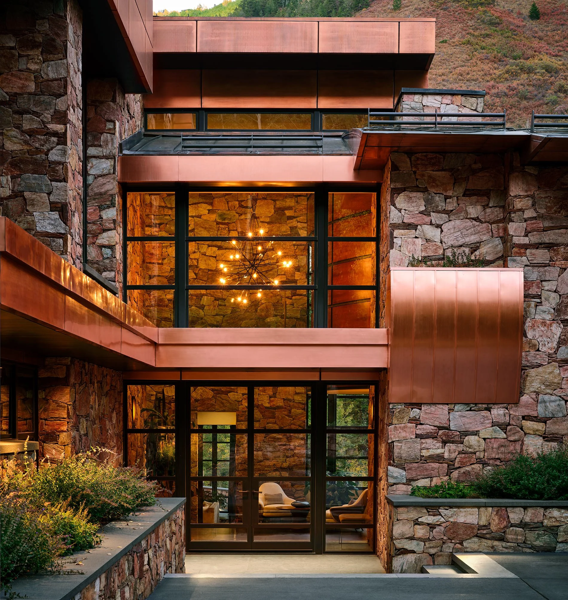 Modern house with stone walls, large glass windows, and copper accents, surrounded by a mountainous landscape.