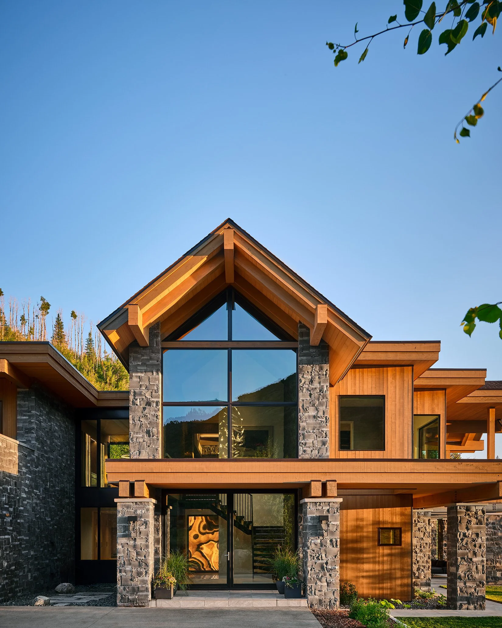 Modern house with large glass windows, wooden and stone exterior, and a gabled roof, surrounded by a landscaped yard and trees against a clear blue sky.