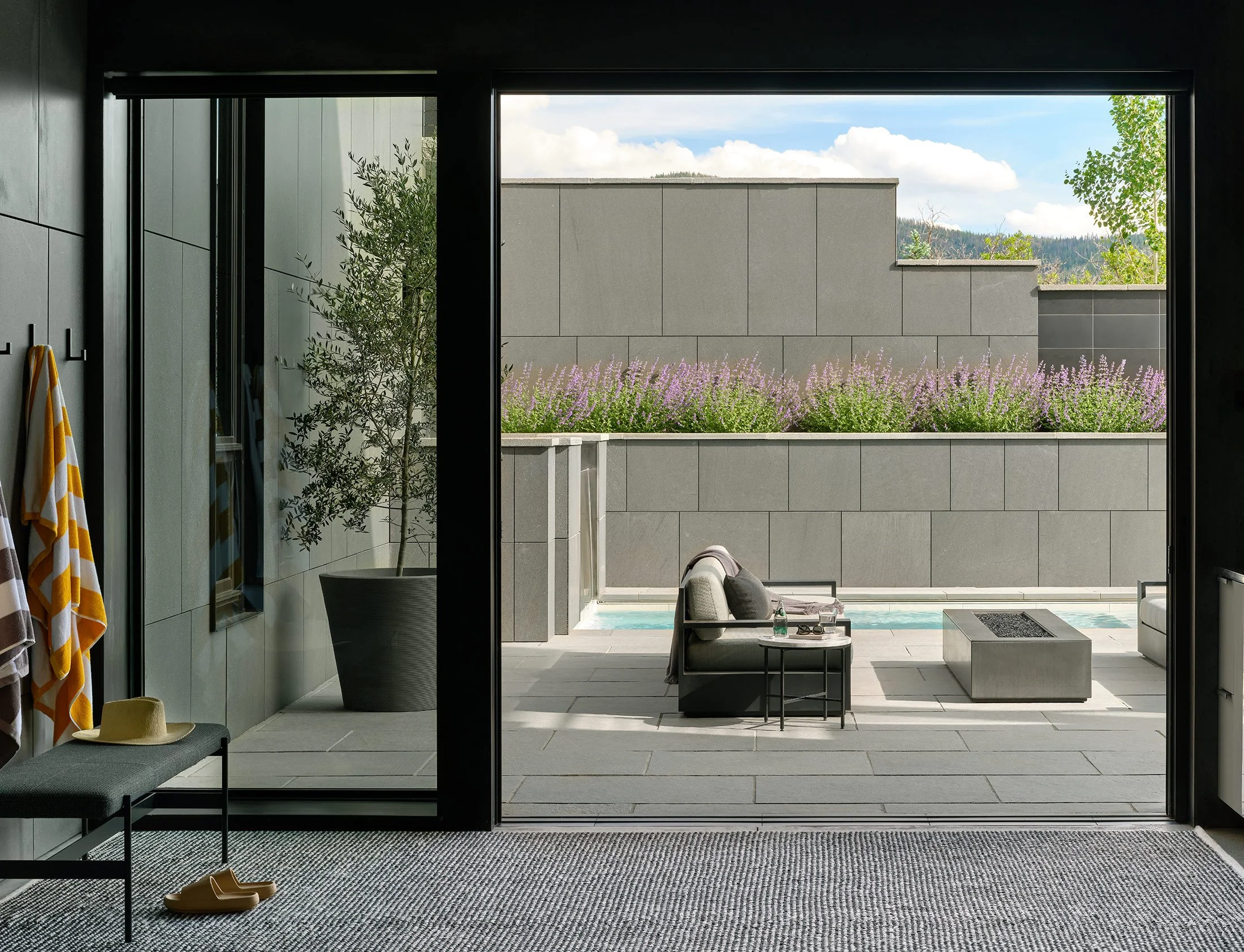 Modern outdoor patio with gray walls, purple flowering plants, a gray bench, armchair, and fire pit, viewed through a large open sliding glass door from an interior room with towels and shoes.