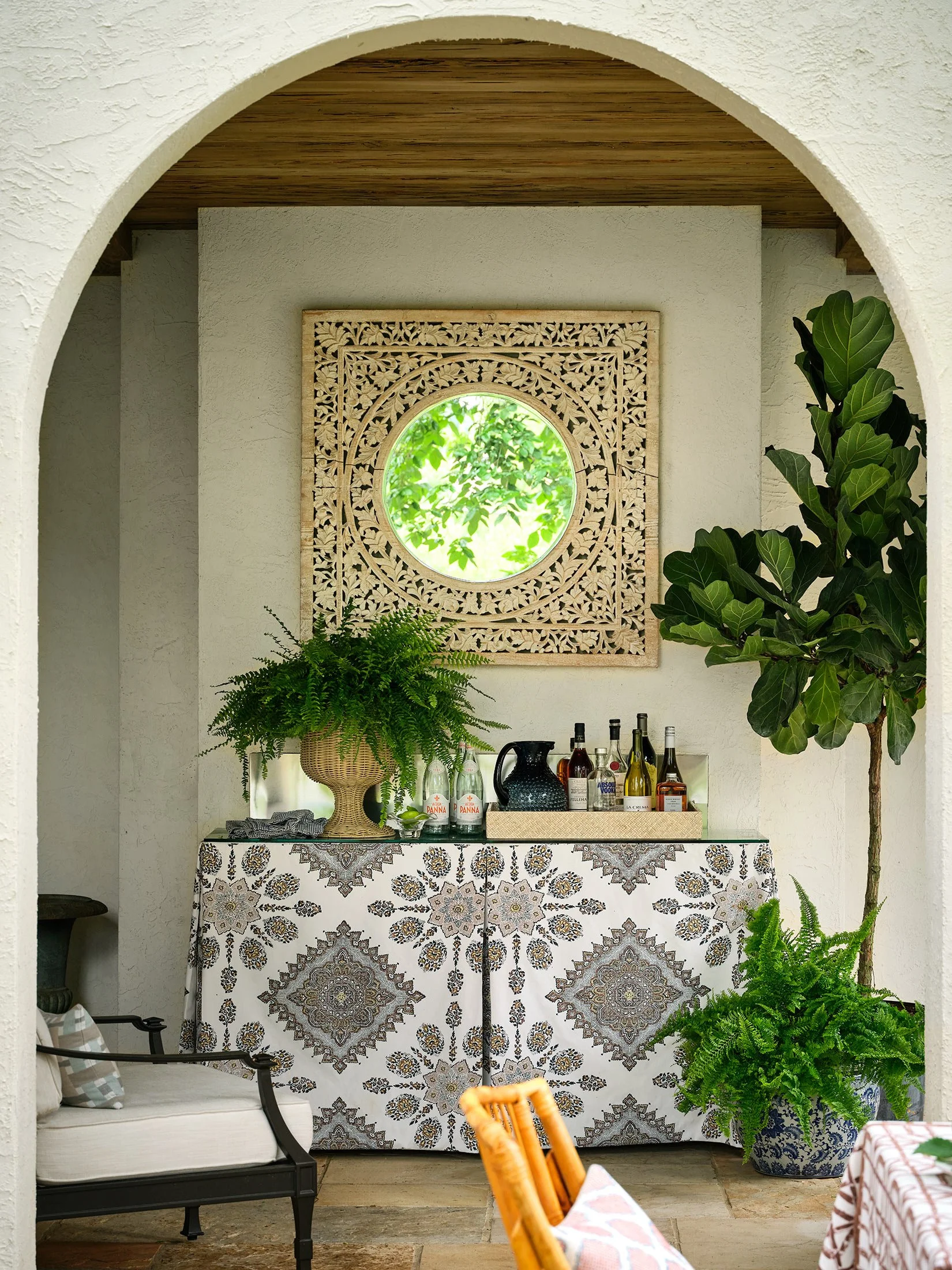 A home bar area with a decorative cloth-covered cabinet, potted plants, bottles of alcohol, and a mirror with a carved wooden frame.