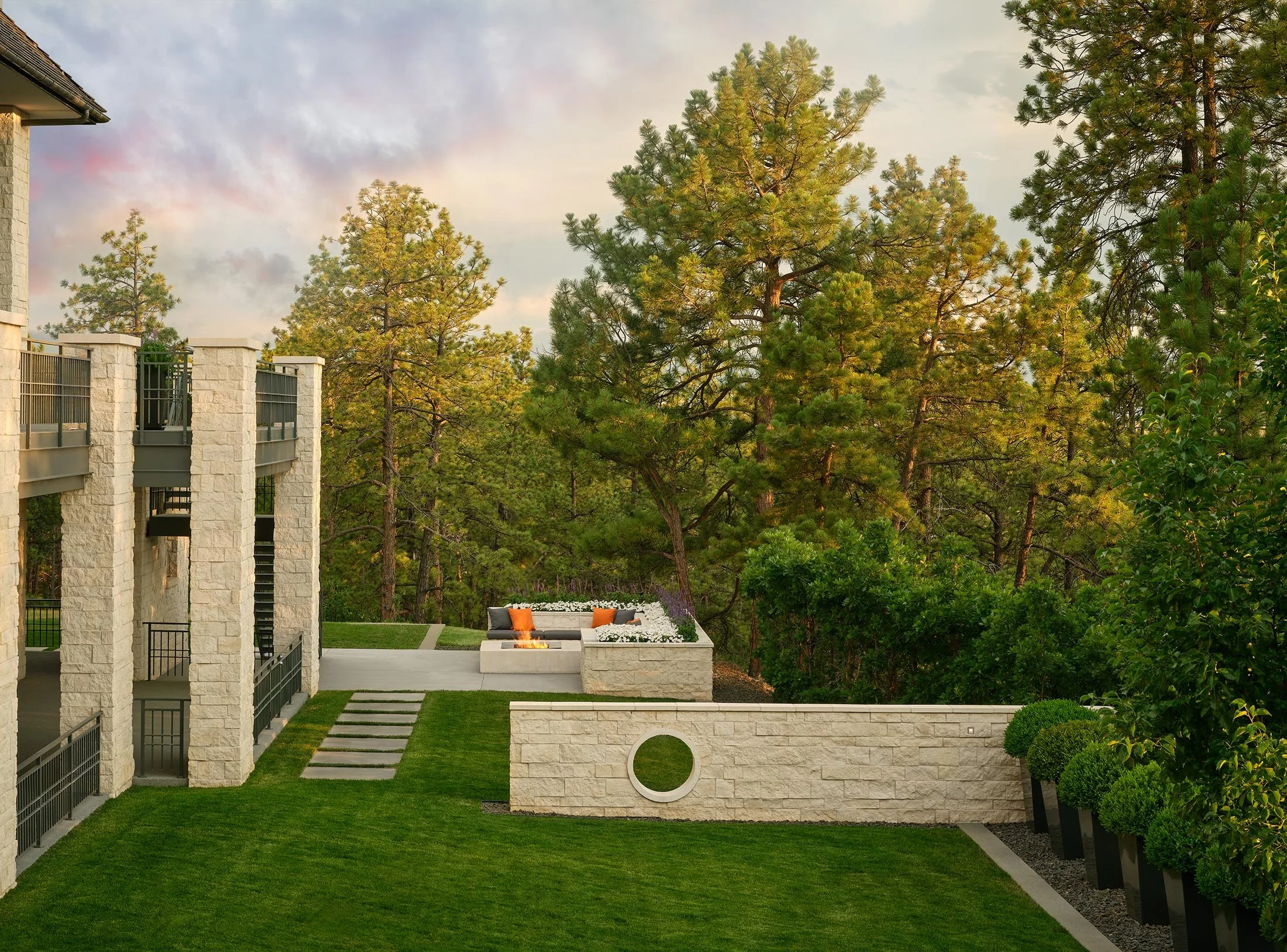 Modern outdoor patio with a fire pit, seating area with orange pillows, surrounded by lush green trees and a manicured lawn.