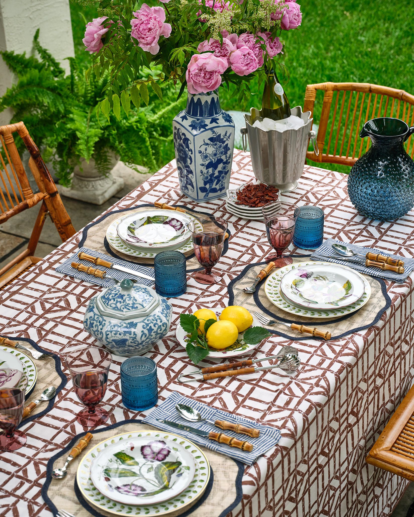 Outdoor dining table set with floral tableware, colorful glassware, a bowl of lemons, a blue and white vase with pink peonies, a silver ice bucket with a wine bottle, and a black textured vase, surrounded by wooden chairs and green foliage.