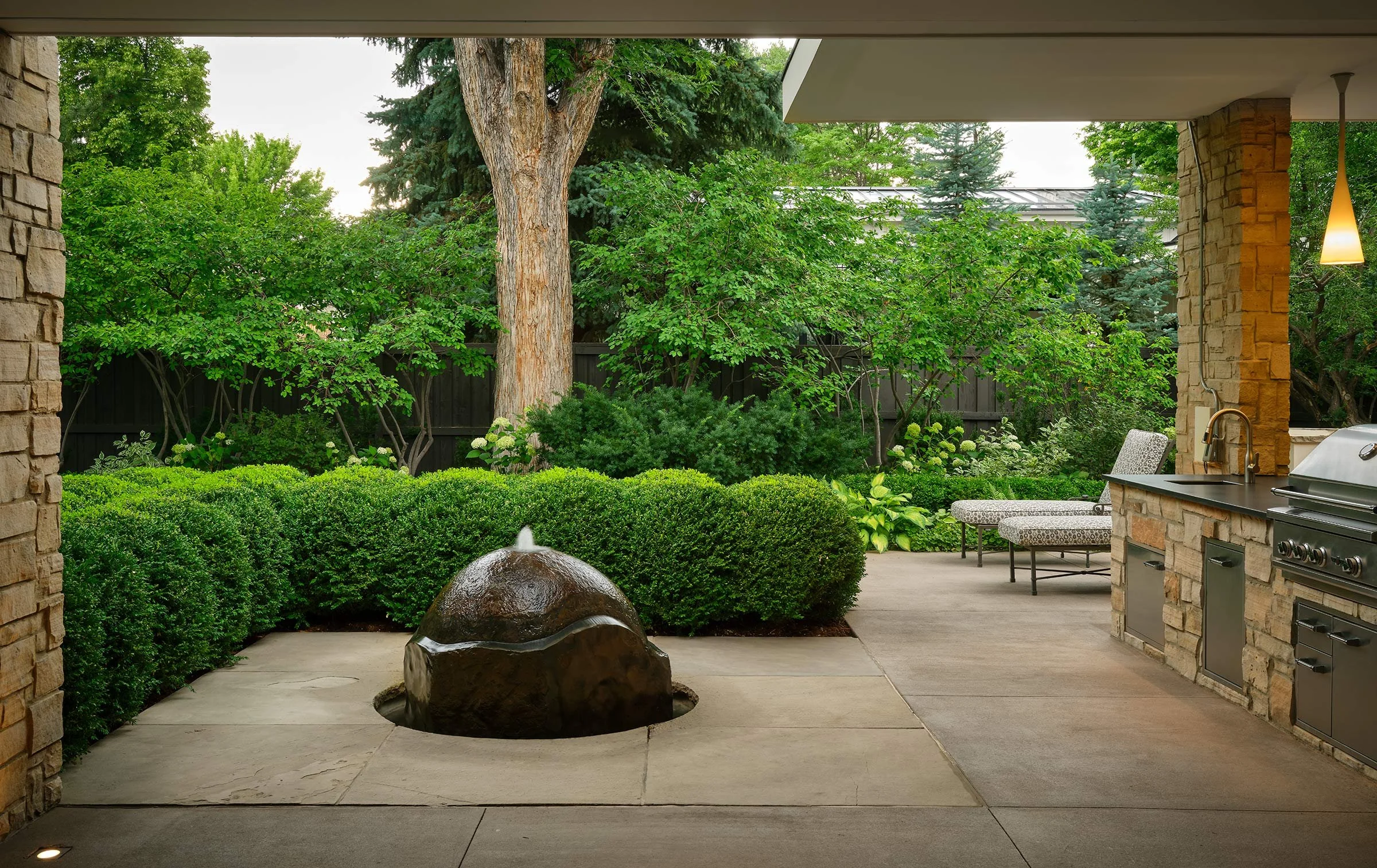 Outdoor patio with lush green trees and shrubs, stone and brick accents, a water fountain, and patio furniture with a built-in grill area.