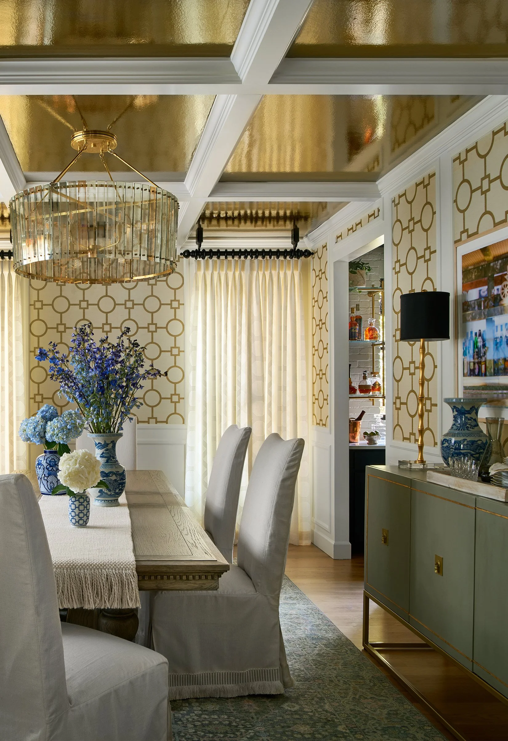 A dining room with a wooden table, beige upholstered chairs, floral arrangements in blue and white vases, gold and glass chandelier, patterned wallpaper, and a sideboard with decorative items.
