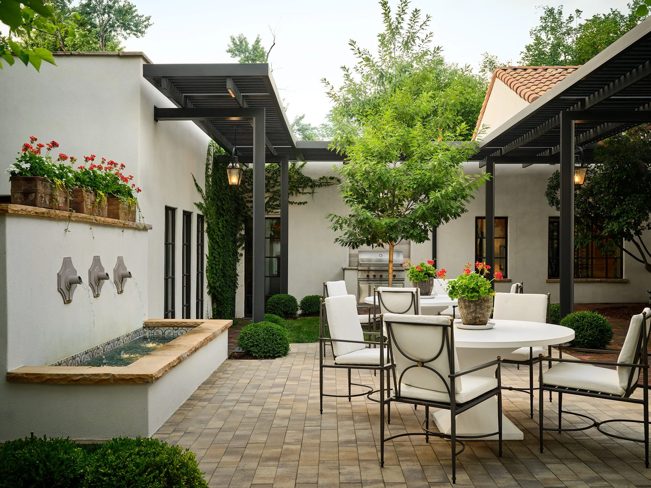 Outdoor patio with white tables, chairs, potted plants, trees, and decorative fountain.