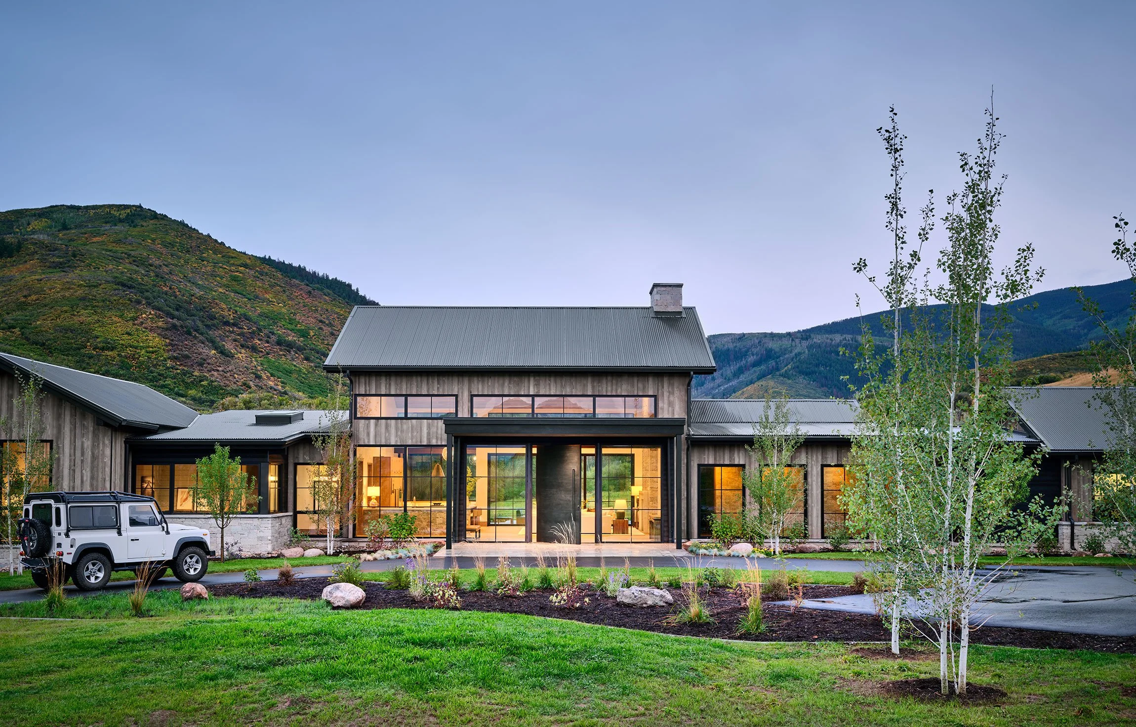 Modern house with metal siding and large glass windows, landscape with green grass, small trees, and mountains in the background at dusk.