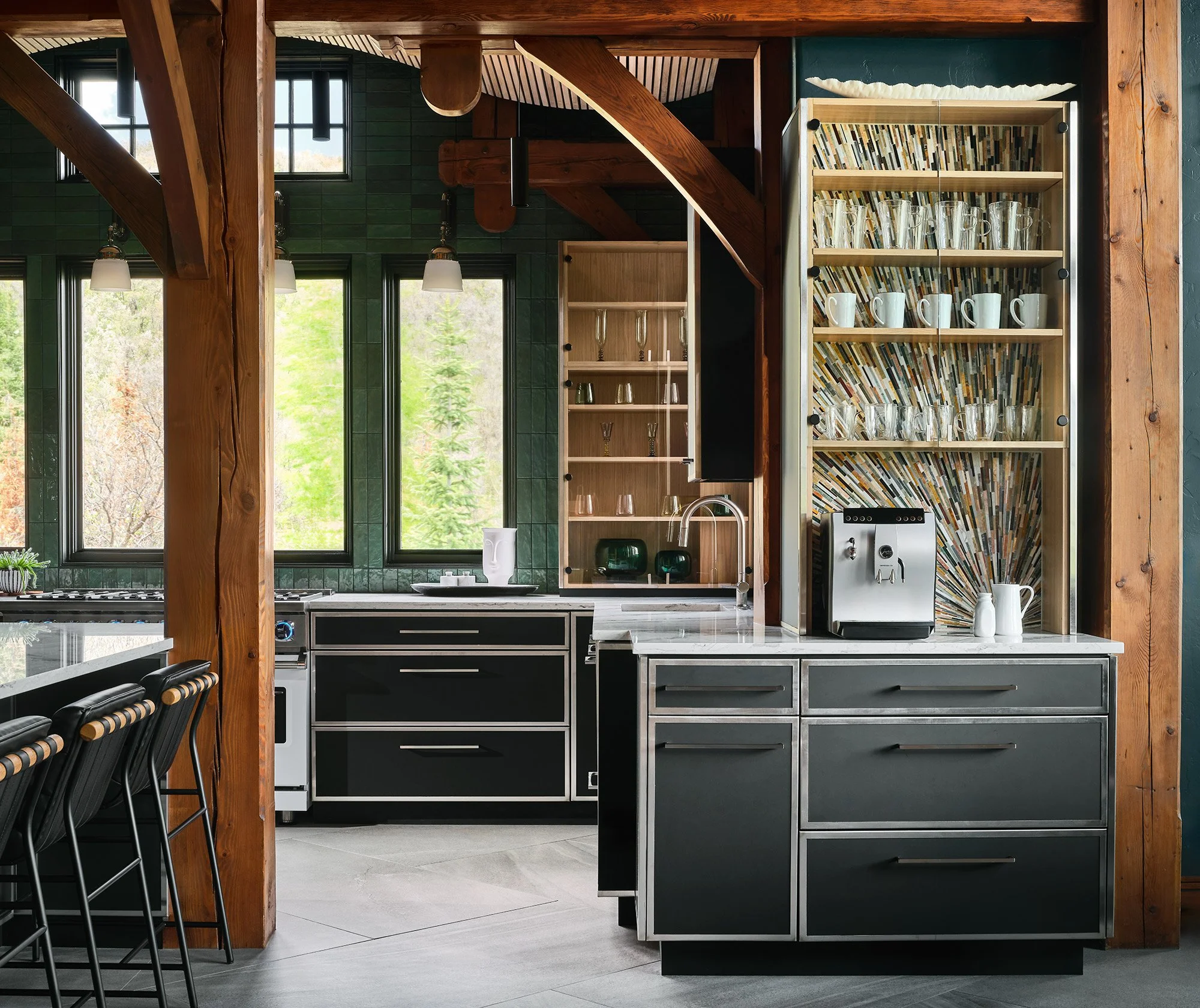 Modern kitchen with green tiled walls, large windows, black and silver cabinets, wooden accents, glassware, and a coffee machine.
