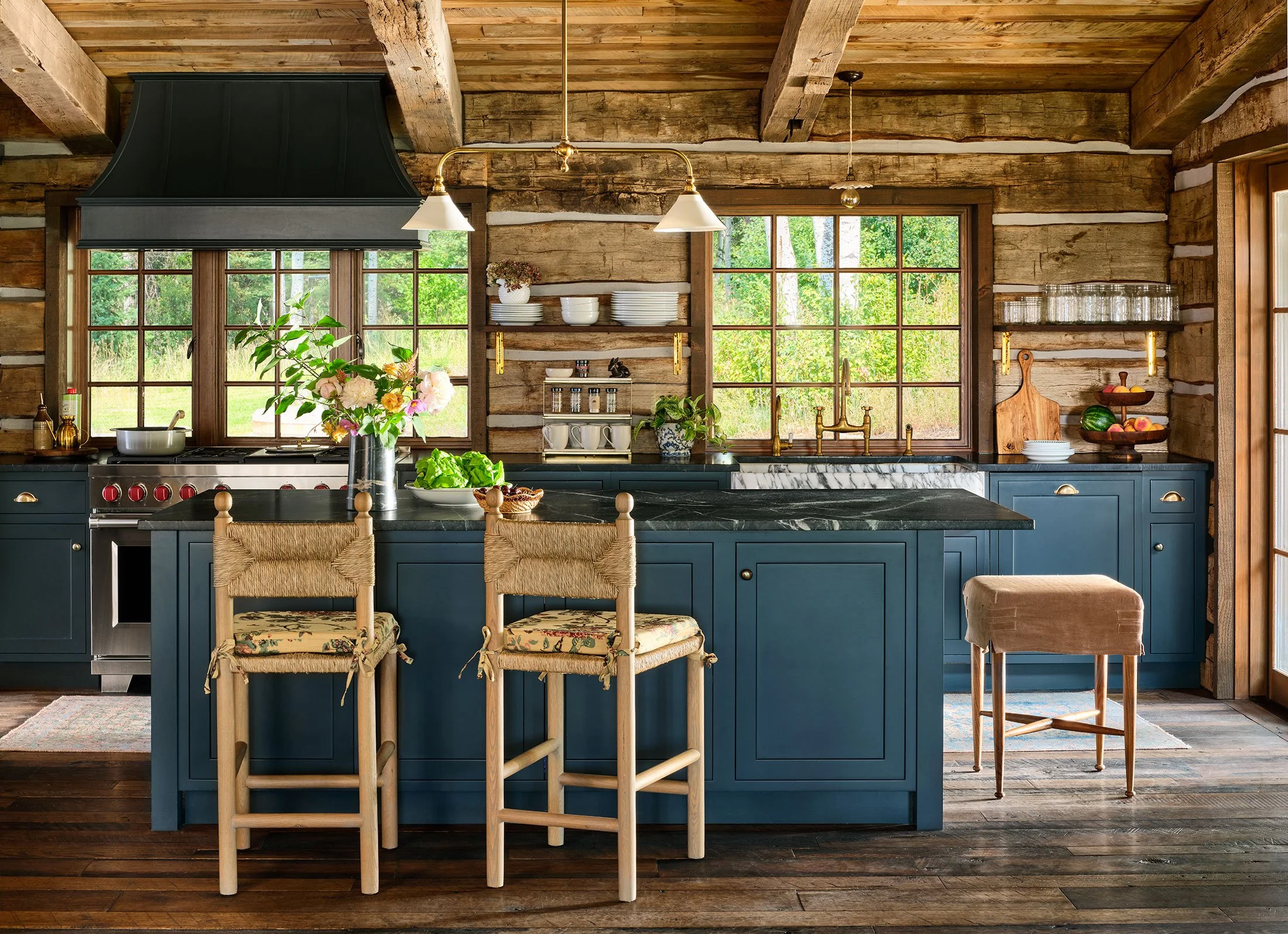 Rustic kitchen with blue cabinets, wooden walls, large windows, black marble countertop, floral centerpiece, bar stools with woven seats, wooden cutting boards, and various kitchenware and decor.