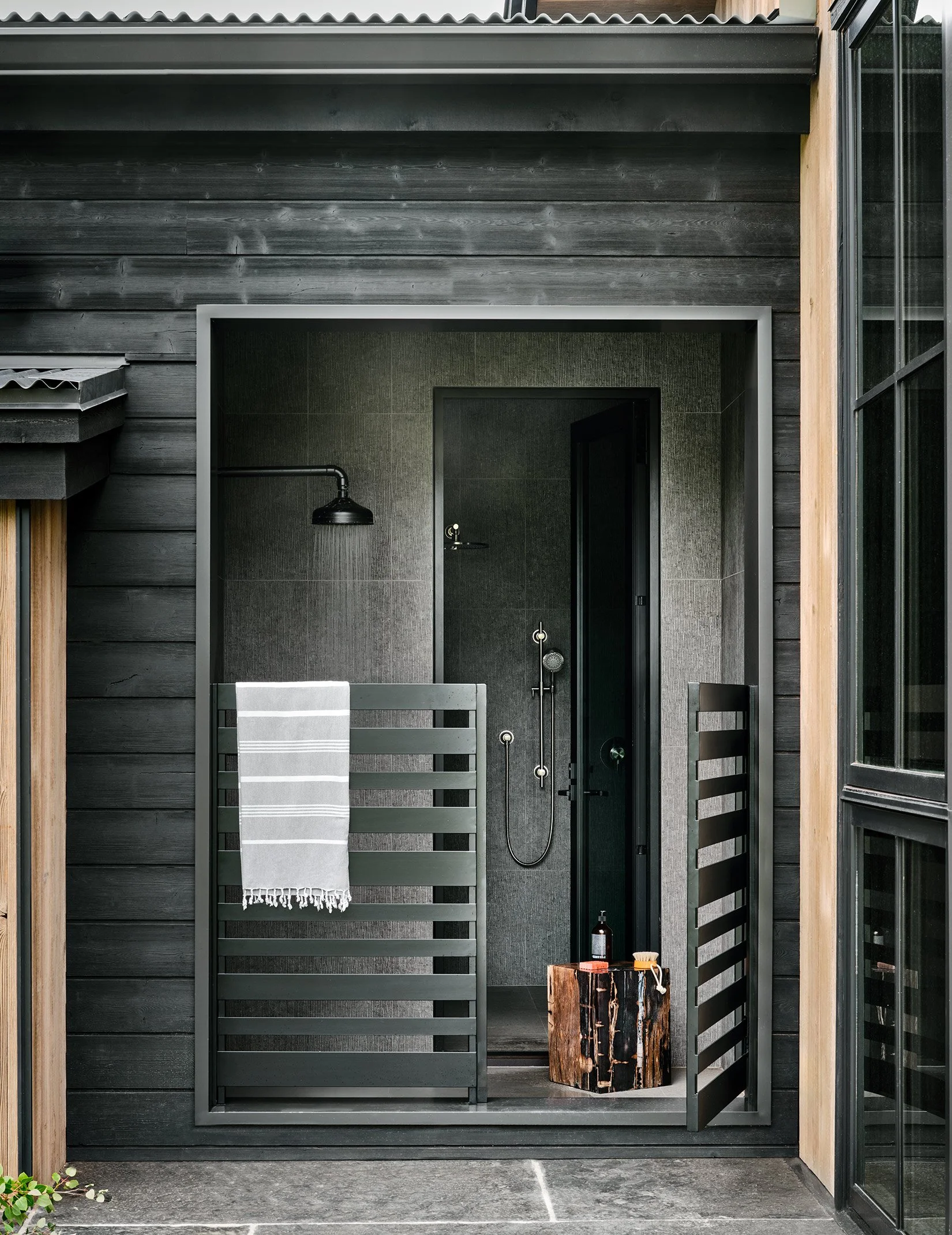 Outdoor shower area with black tiles, a rain showerhead, a towel hanging on a black wooden privacy screen, a small wooden stool with bath items, and a black door.