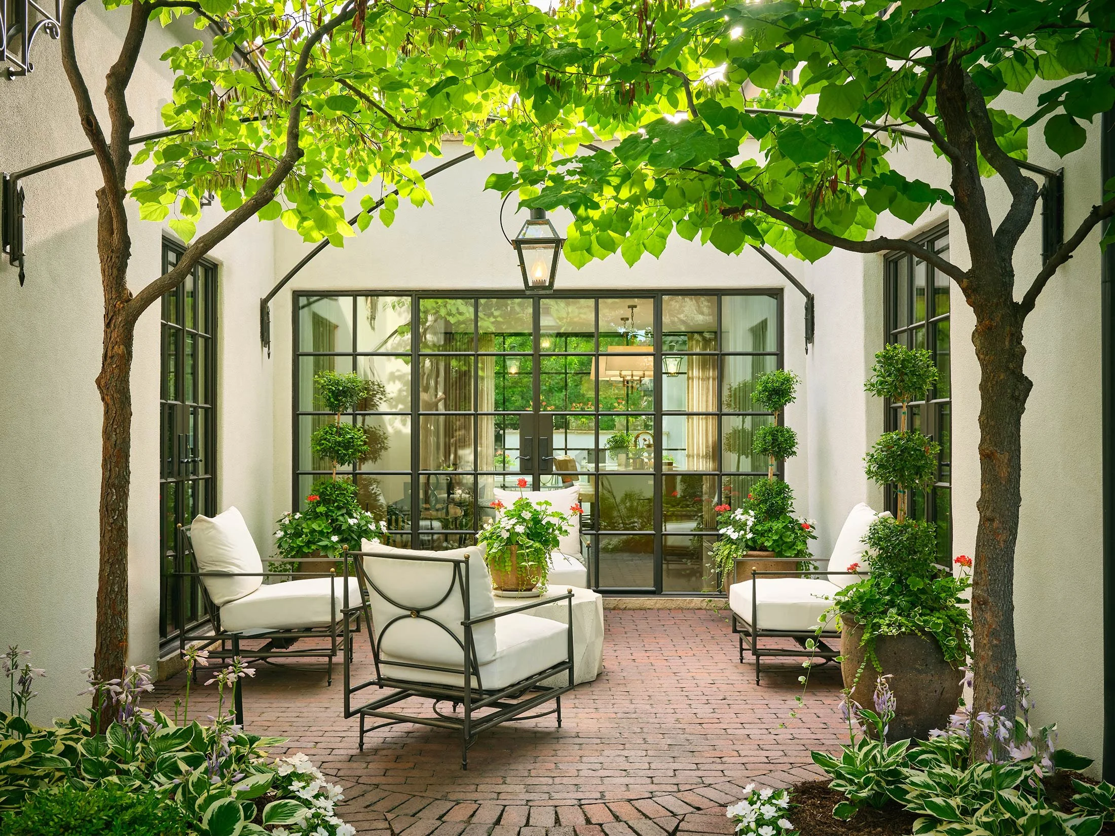 A cozy outdoor patio with white cushioned chairs, potted plants, and lush greenery, enclosed by black metal framed glass doors and windows.