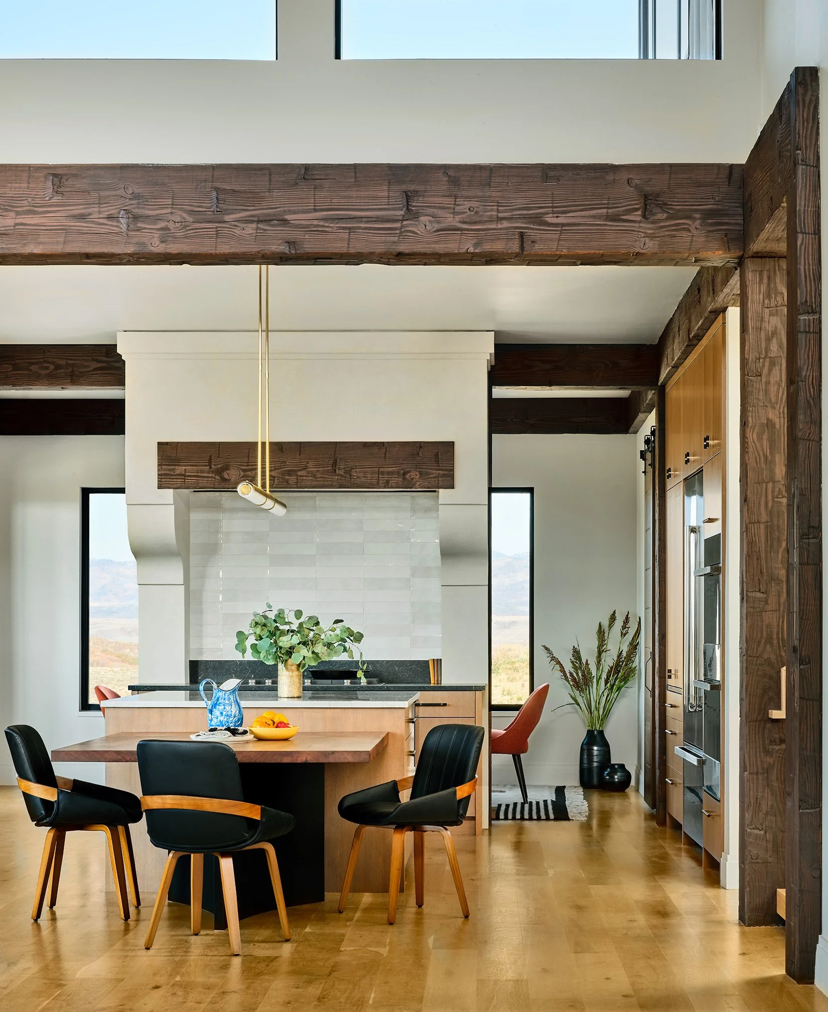 A modern kitchen with wooden beams, a round wooden dining table, four black chairs with wooden accents, and large windows showing a mountain view.