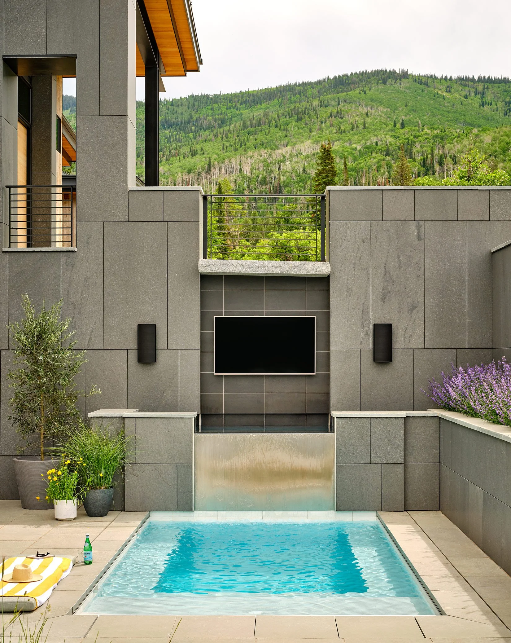 Modern outdoor space featuring a small swimming pool with a waterfall, gray stone walls, potted plants, a wall-mounted TV, and a mountain range in the background.