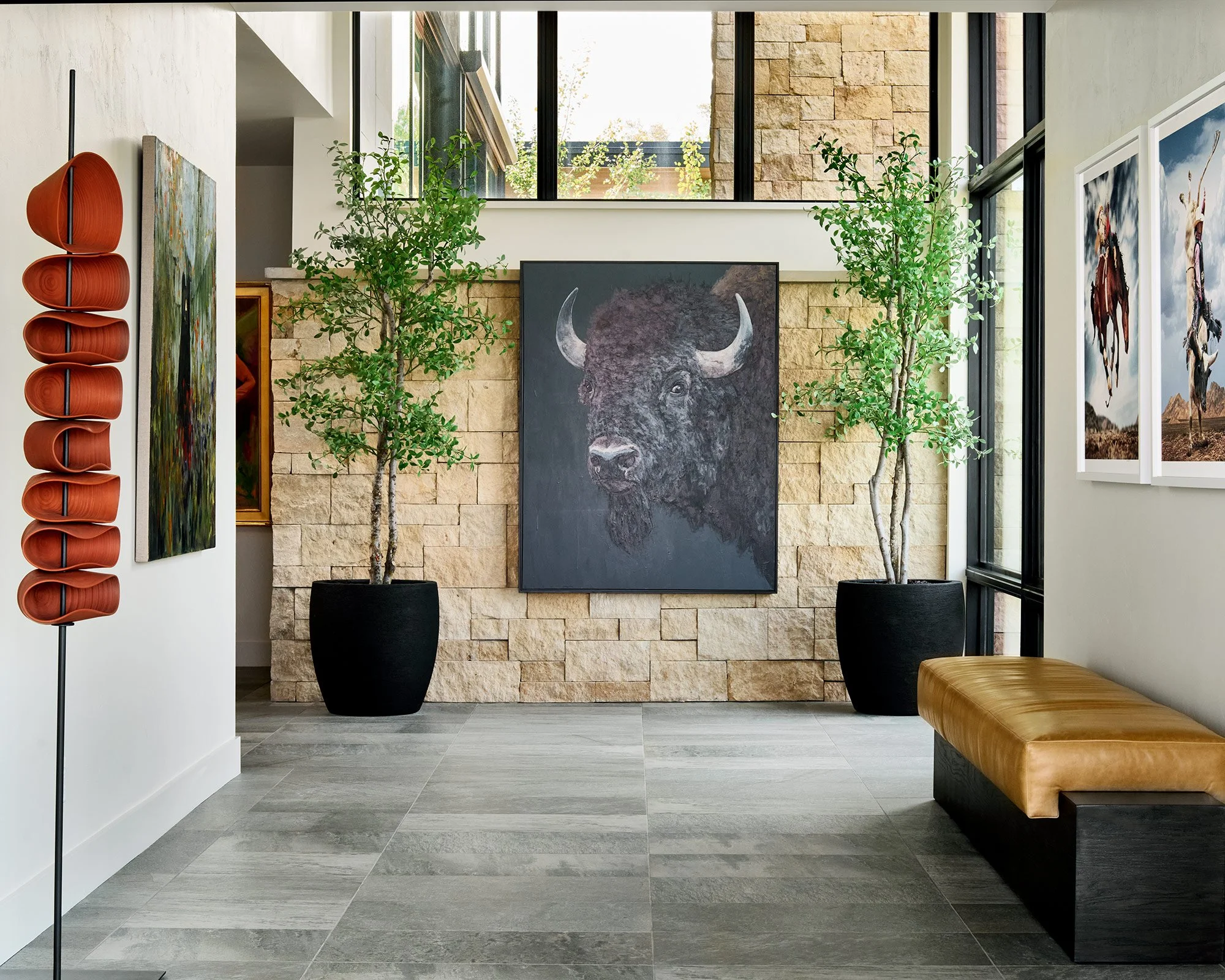 Modern interior lobby with stone accent wall, large buffalo portrait, two potted trees, colorful artwork, and a yellow bench.