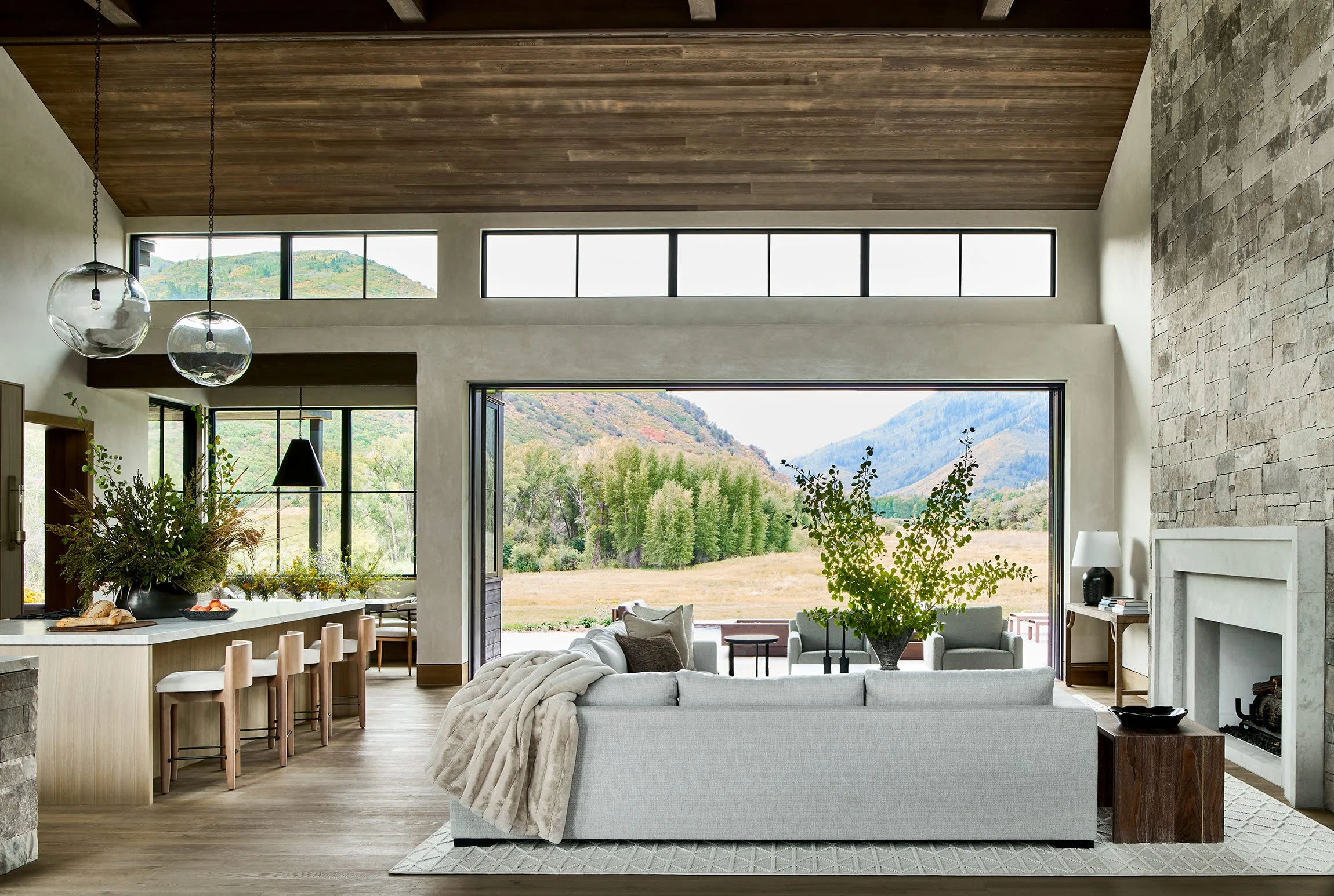 Living room with large open glass doors revealing a scenic mountain view, modern sofa with throw blanket, stone fireplace, and wooden ceiling.