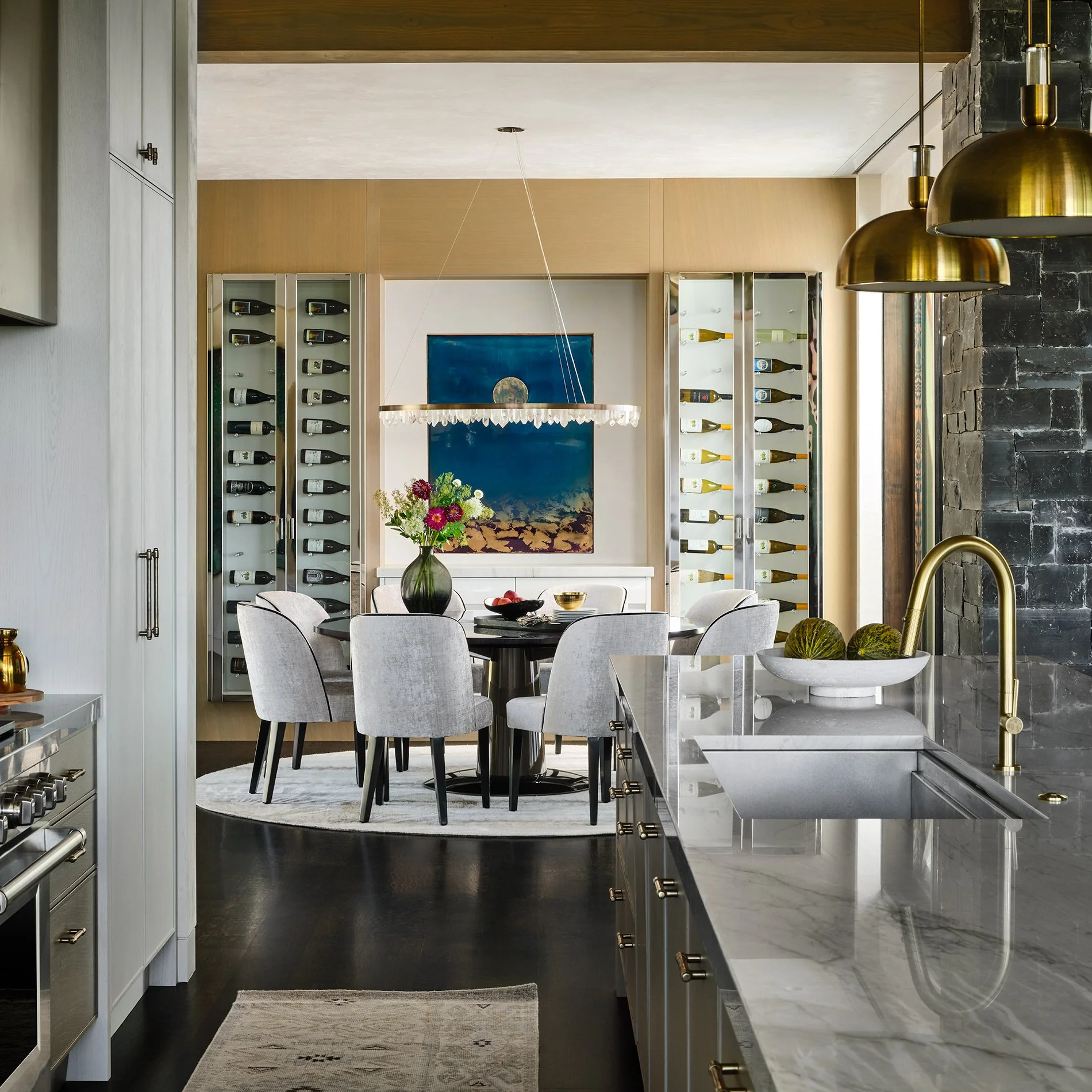 Modern dining area with a round table, six gray upholstered chairs, a vase of flowers, and wall-mounted wine racks with bottles. The room has a large abstract painting, a crystal chandelier, and a marble kitchen counter with a gold faucet.